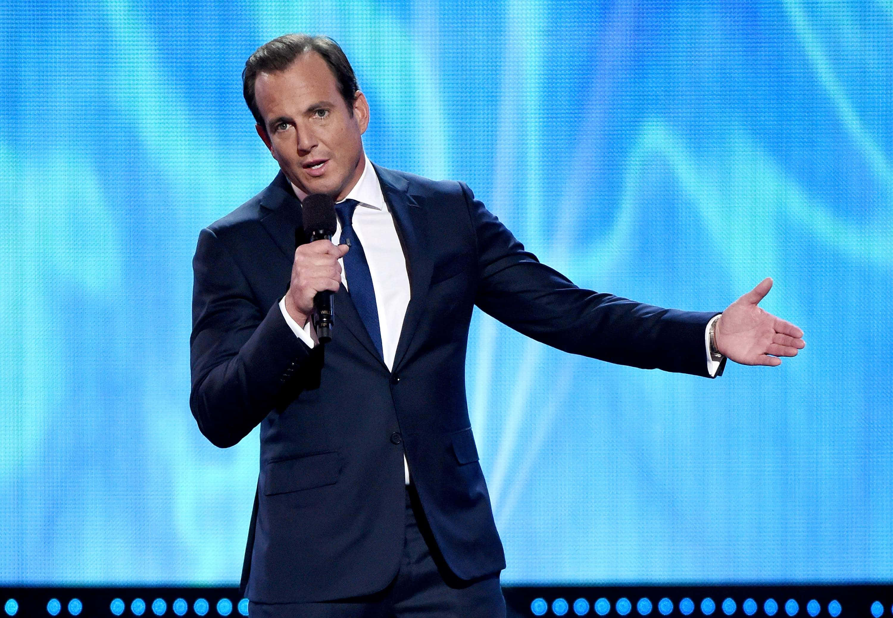 Will Arnett 桌布 壁紙 — 一名身穿深色西裝的男子手持麥克風，在充滿活力的藍色數位背景前熱情地做手勢。