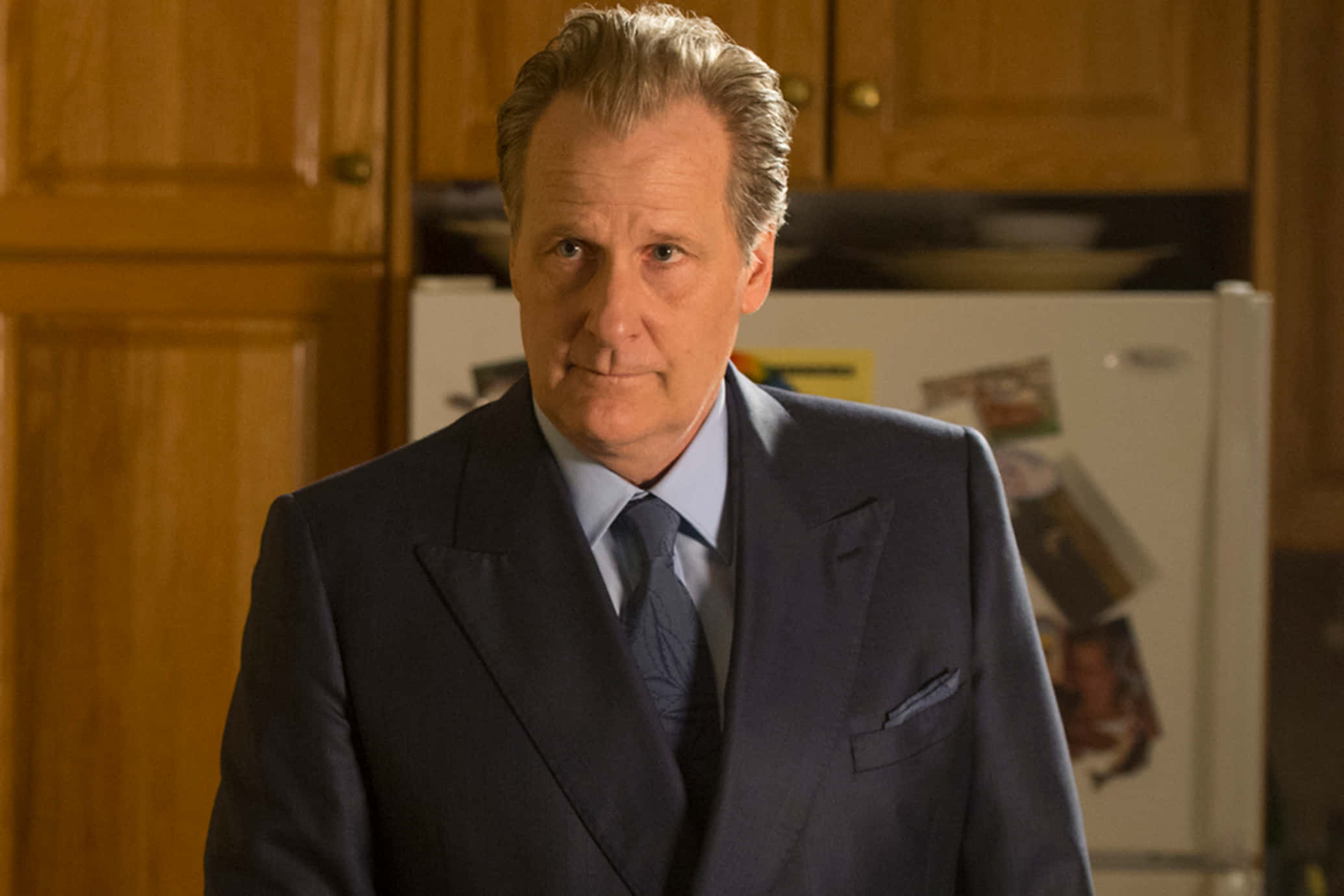 Jeff Daniels 桌布 壁紙 — 一名身穿深色西裝的男子站在一個燈光溫暖的廚房裡，身後有木製櫥櫃和一個貼滿照片的冰箱。