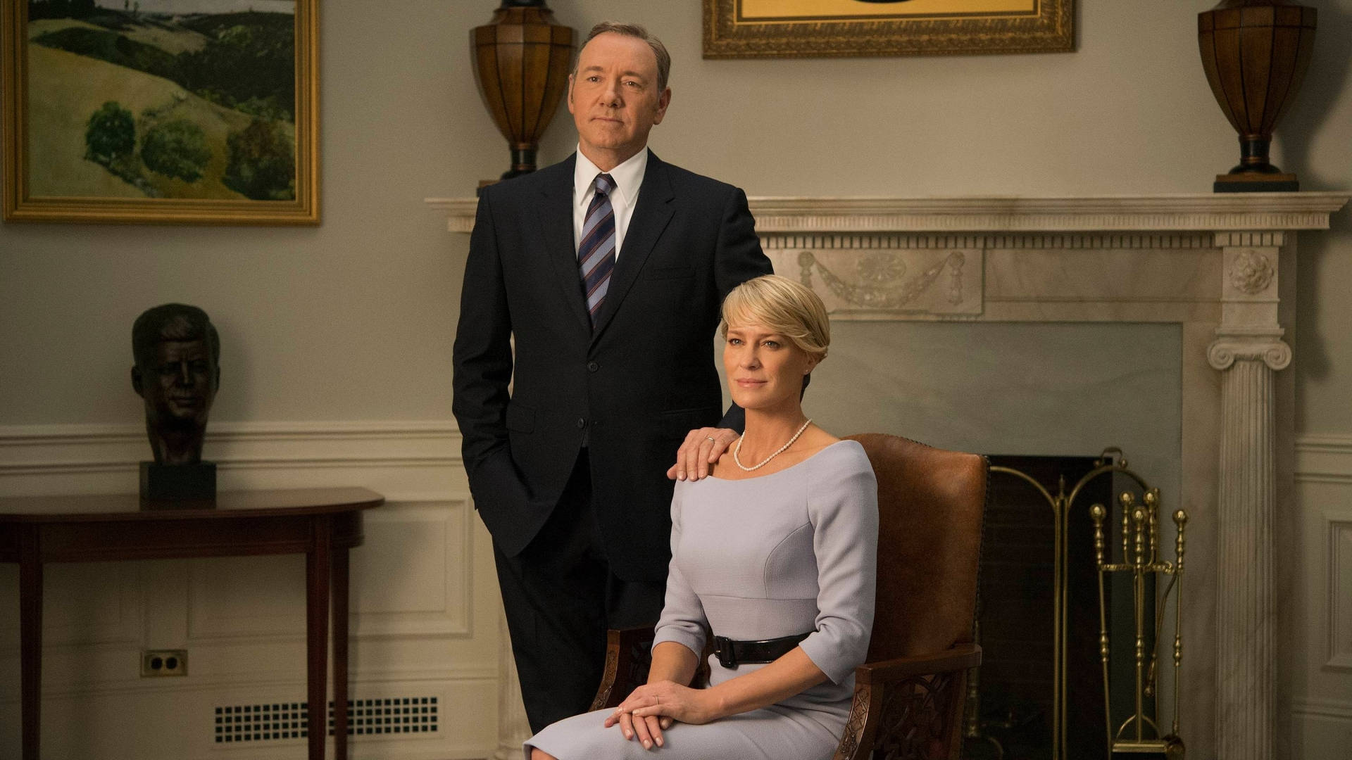 kevin spacey 桌布 壁紙 — 一名身穿深色西裝的男子站在一名身穿淺灰色連衣裙的坐著女子身後，位於一個帶有壁爐和裱框畫作的正式辦公室內。