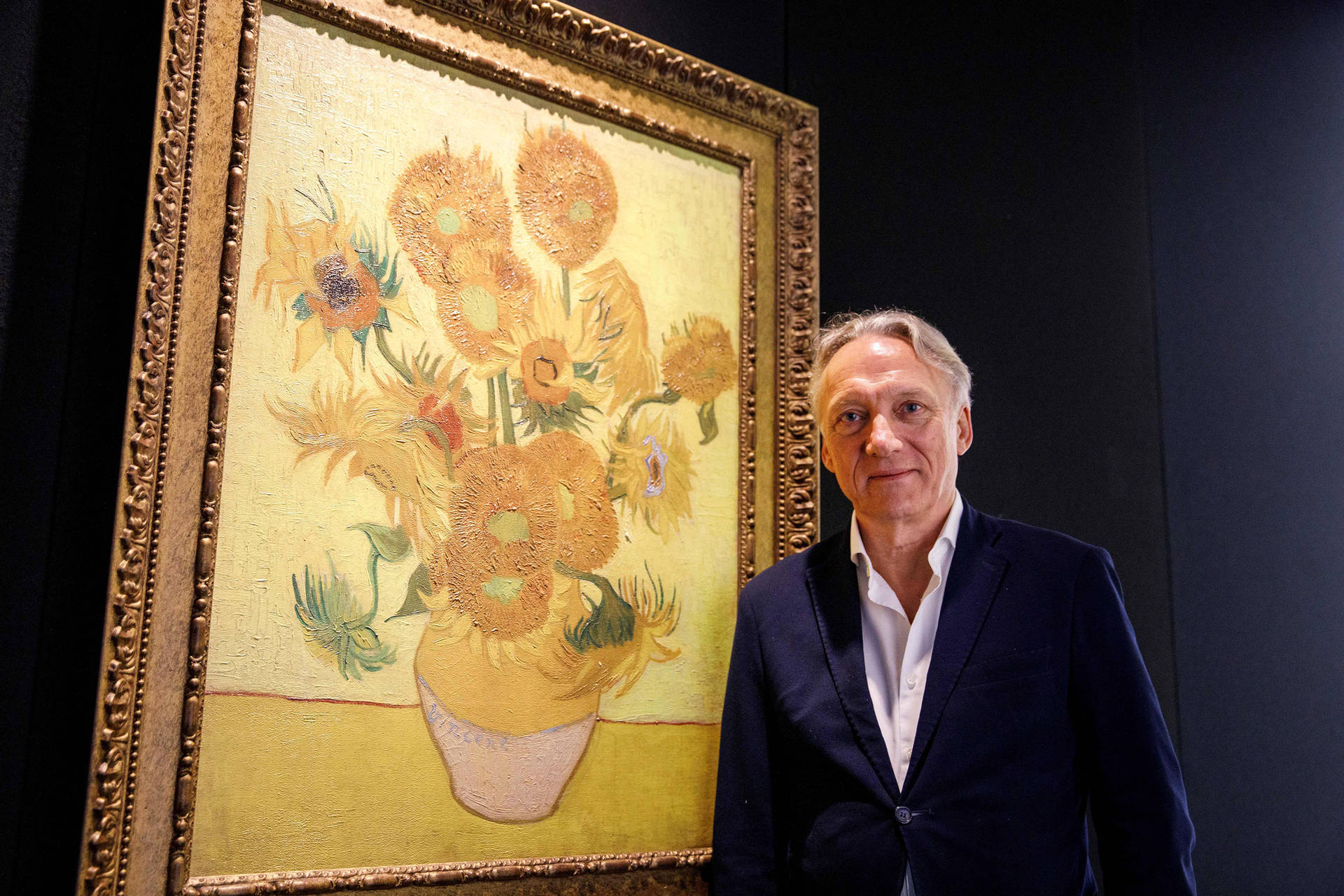 Museum 桌布 壁紙 — 一名身穿深色西裝的男子站在昏暗畫廊中一幅梵谷的大型向日葵畫框旁邊。
