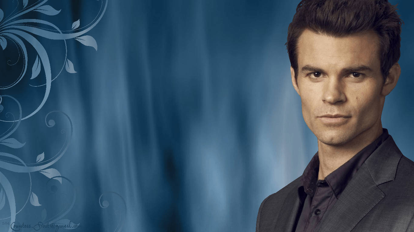 elijah mikaelson 桌布 壁紙 — 一名身穿深色西裝的男子站在藍色窗簾背景前，左側有華麗的白色花卉漩渦圖案。