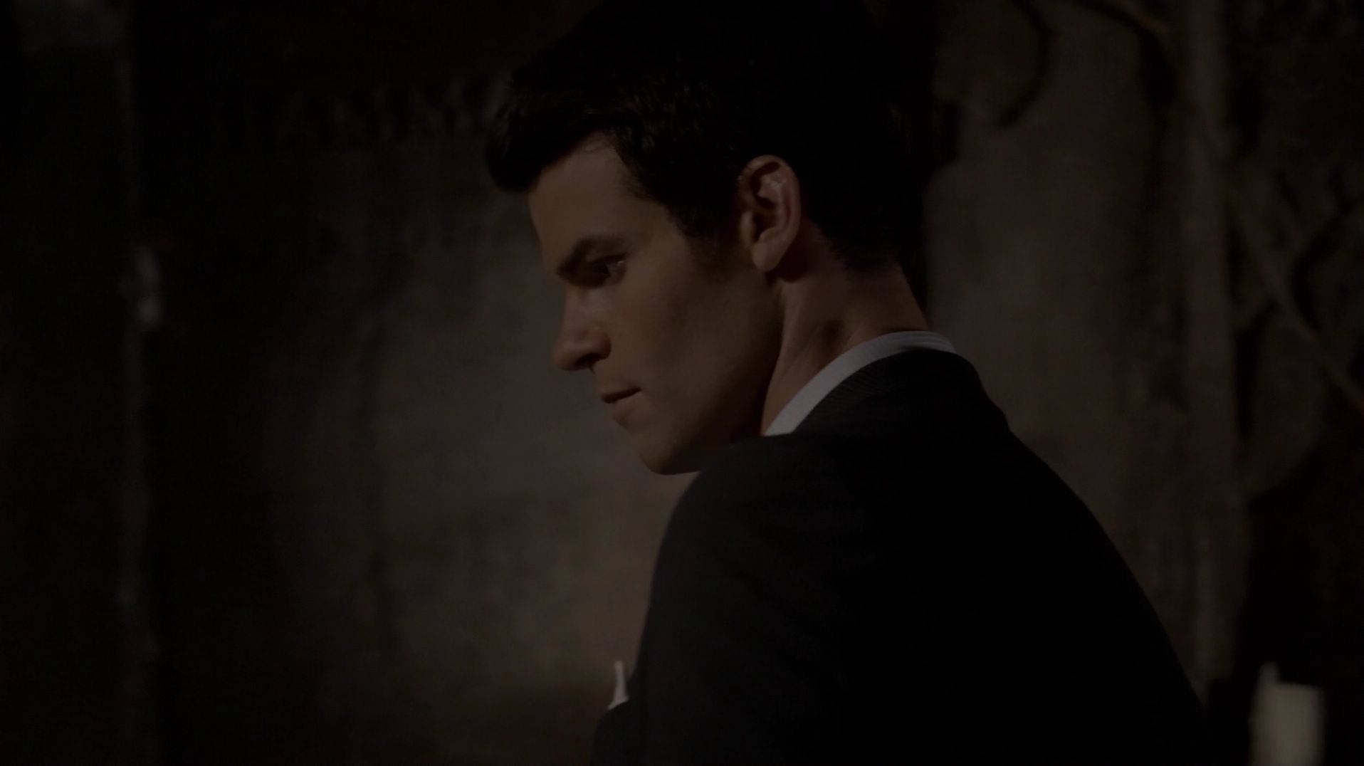 elijah mikaelson 桌布 壁紙 — 一名身穿深色西裝的男子站在陰影昏暗處，表情嚴肅，微弱的光線勾勒出他的側面輪廓。