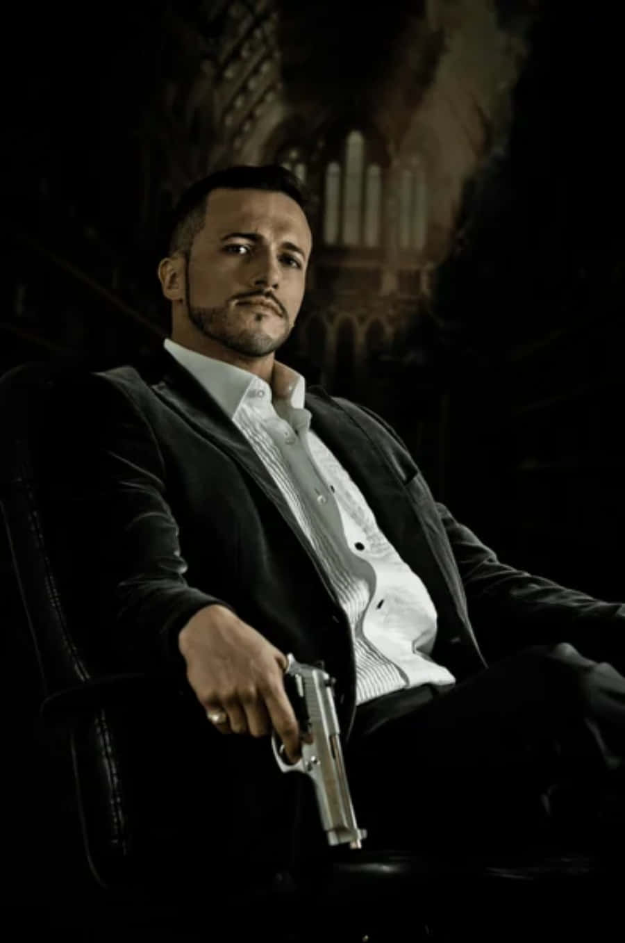 Mafia 桌布 壁紙 — 一名身穿深色西裝的男子自信地坐在皮椅上，手持銀色手槍，背景是燈光昏暗的哥德式大教堂。
