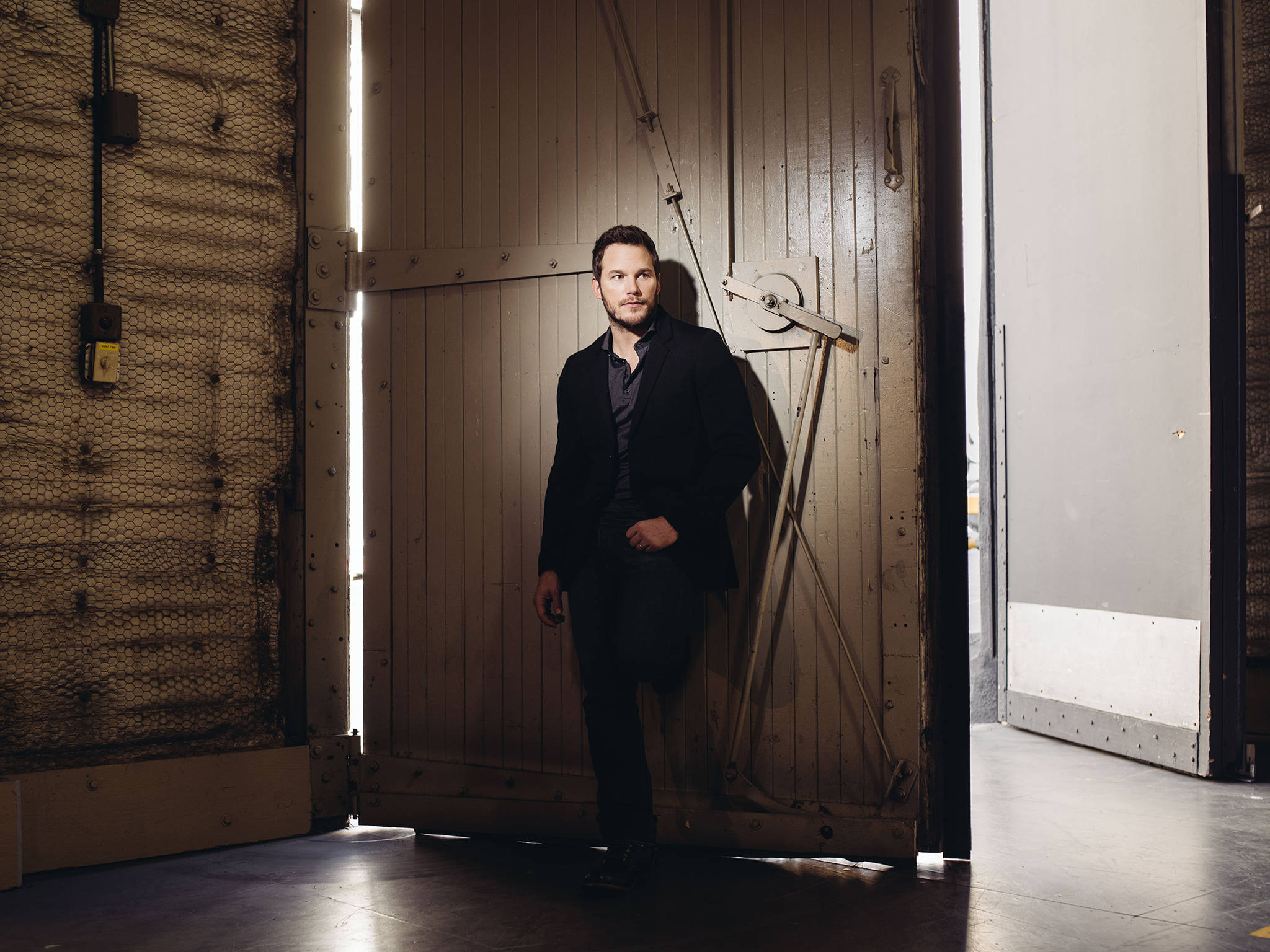 Chris Pratt 桌布 壁紙 — 一名身穿深色西裝的男子靠在倉庫的大型工業門上，側光效果強烈。