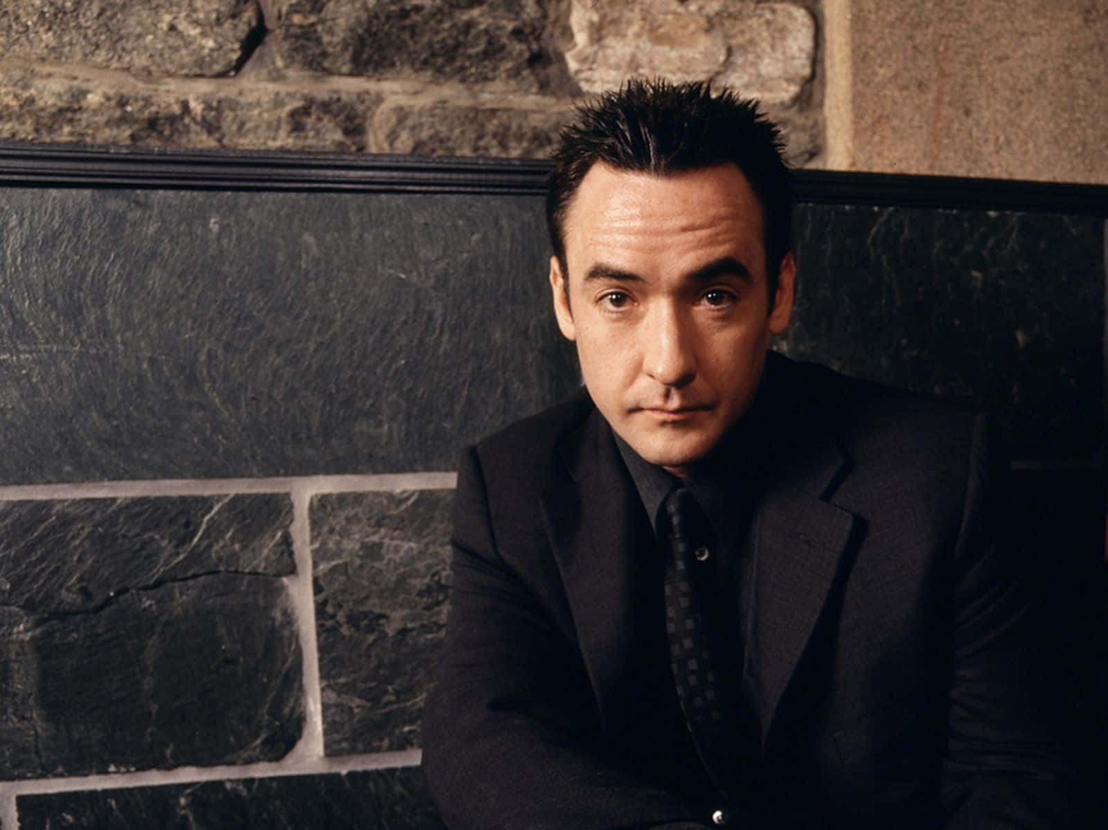 John Cusack 桌布 壁紙 — 一名身穿深色西裝的男子靠著石牆坐下，在戲劇性的燈光下目光專注。