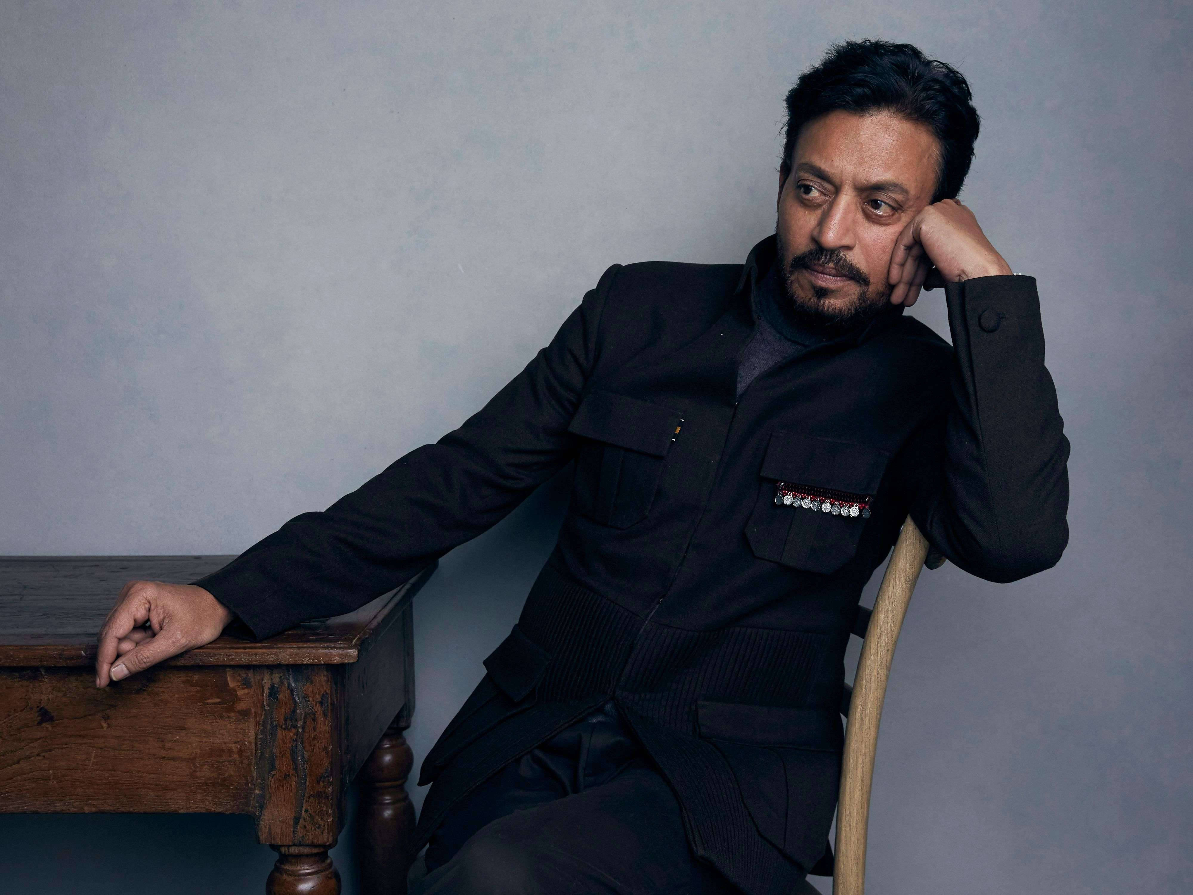 Irrfan Khan 桌布 壁紙 — 一名身穿深色訂製外套的男子，倚靠在一張質樸的木桌上，背景是柔和的灰色牆壁，神情若有所思。