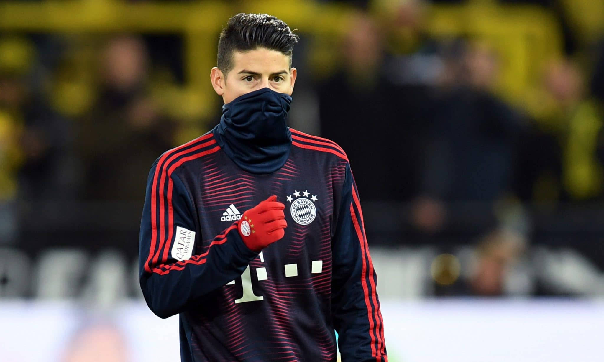 James Rodriguez 桌布 壁紙 — 一名身穿深色訓練服（帶紅色條紋）的足球運動員站在球場上，戴著黑色面罩和紅色手套。