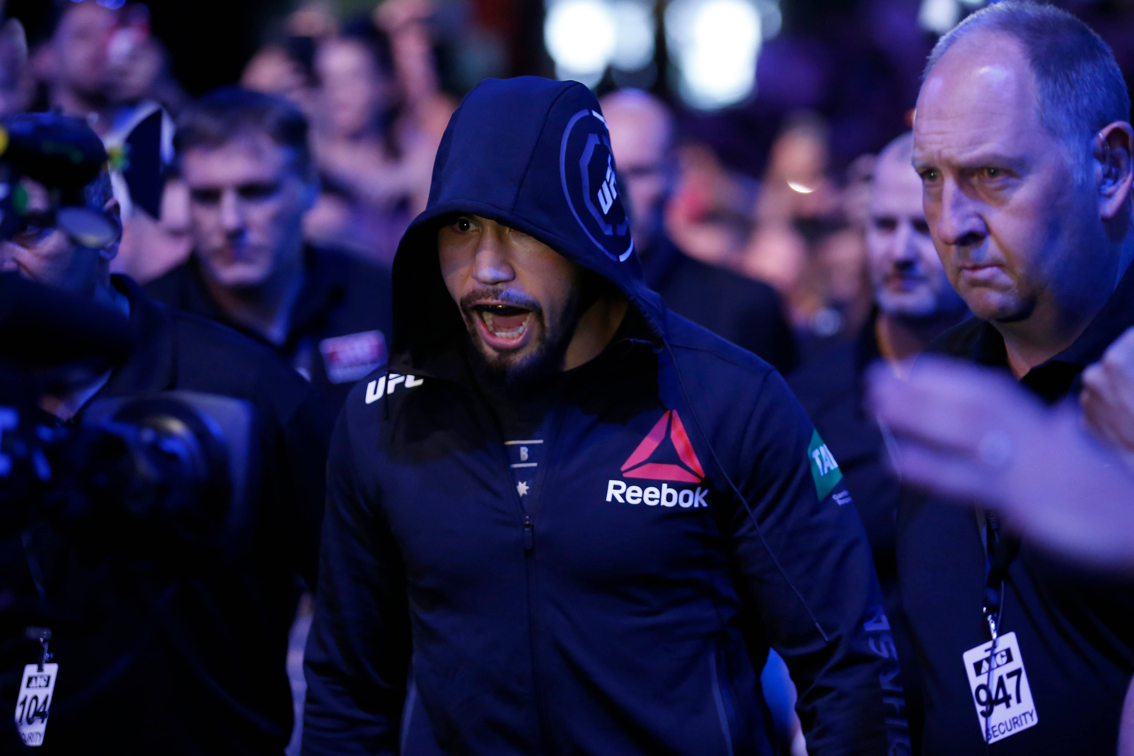 UFC 桌布 壁紙 — 一名身穿深色連帽衫和Reebok外套的戰士在藍色舞台燈光下被保安和攝影機包圍，激烈地大喊。