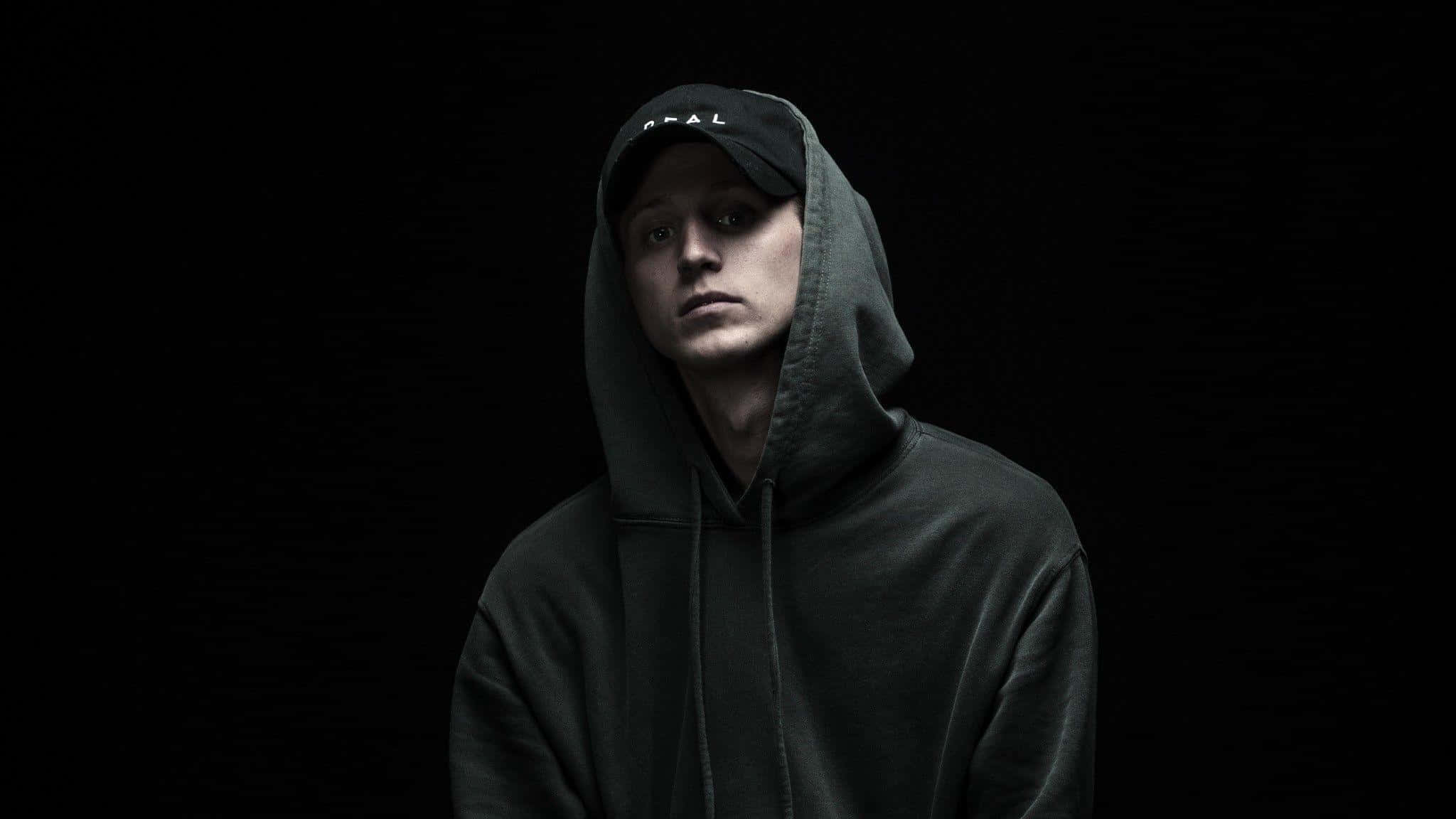 Nf Rapper 桌布 壁紙 — 一名身穿深色連帽衫和帽子的年輕男子在戲劇性的攝影棚燈光下，對著黑色背景專注地凝視。
