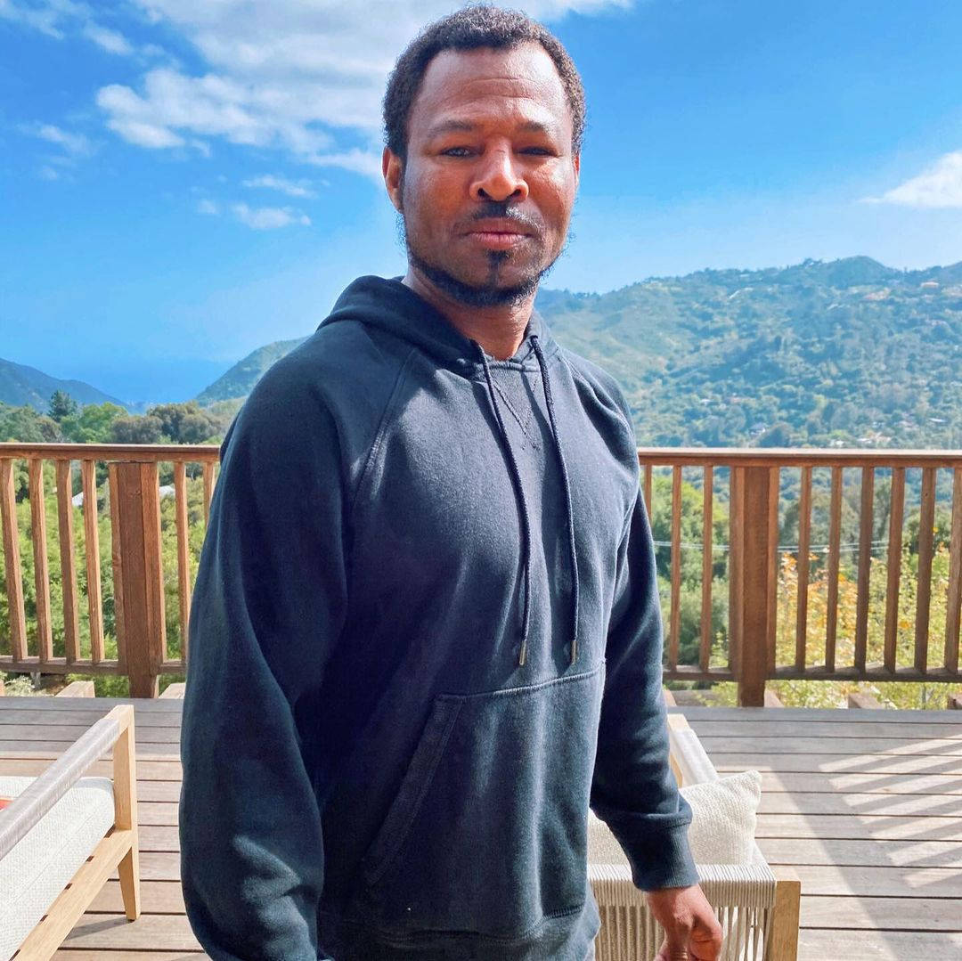 Celebrity 桌布 壁紙 — 一名身穿深色連帽衫的男子站在木製甲板上，背景是明亮的藍天下連綿的山脈和翠綠的山丘。