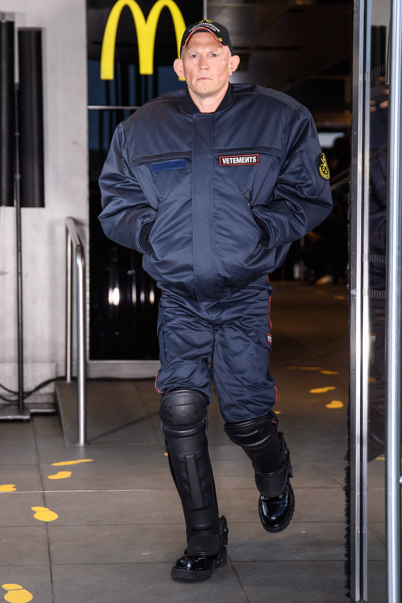 Vetements 桌布 壁紙 — 一名身穿深藍色VETEMENTS連身衣和黑色護膝的男子走過麥當勞入口，地板上有黃色腳印。