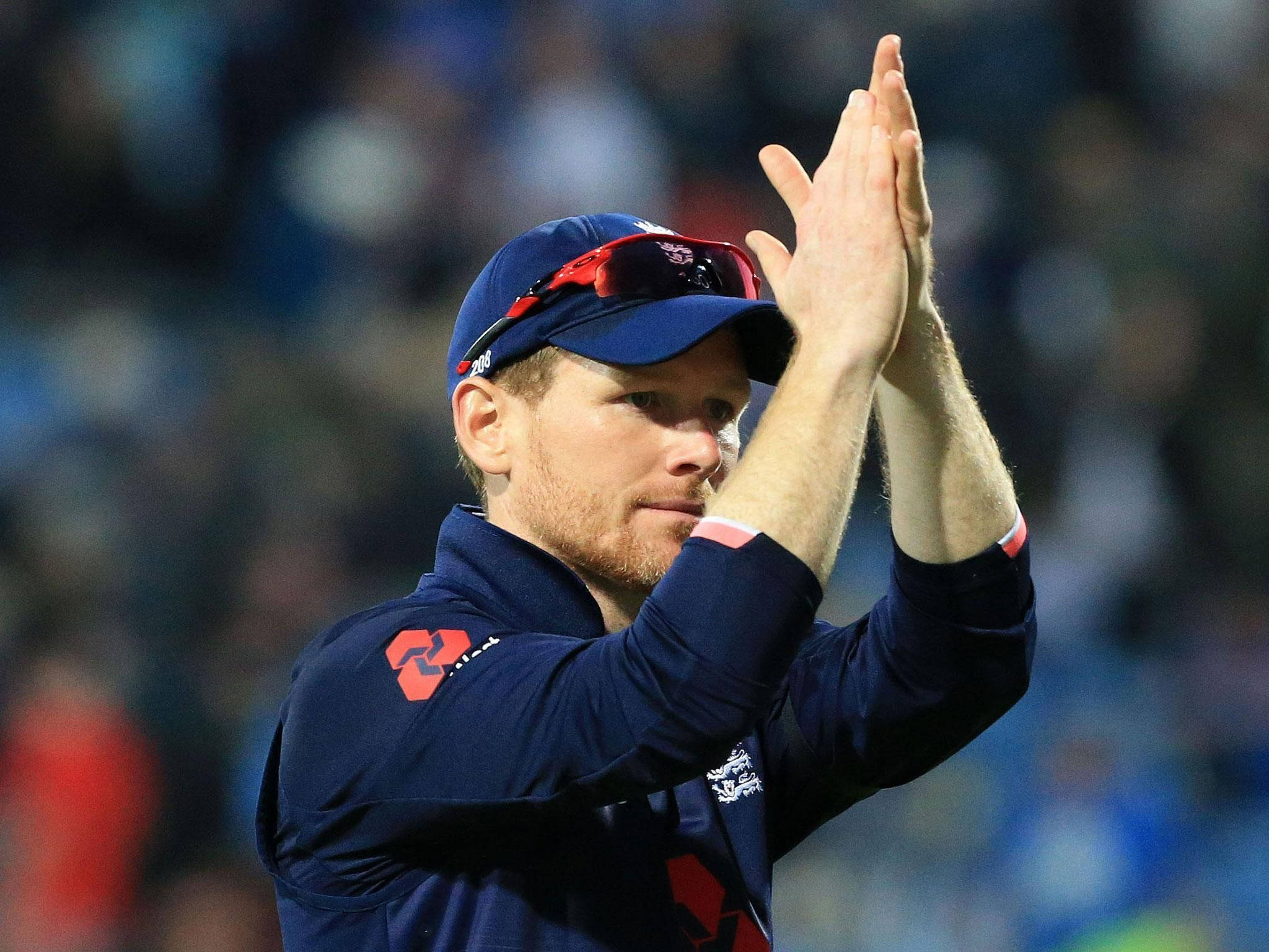 eoin morgan 桌布 壁紙 — 一名身穿深藍色制服和帽子的板球運動員在夜間比賽中，在體育場燈光下鼓掌表示讚賞。
