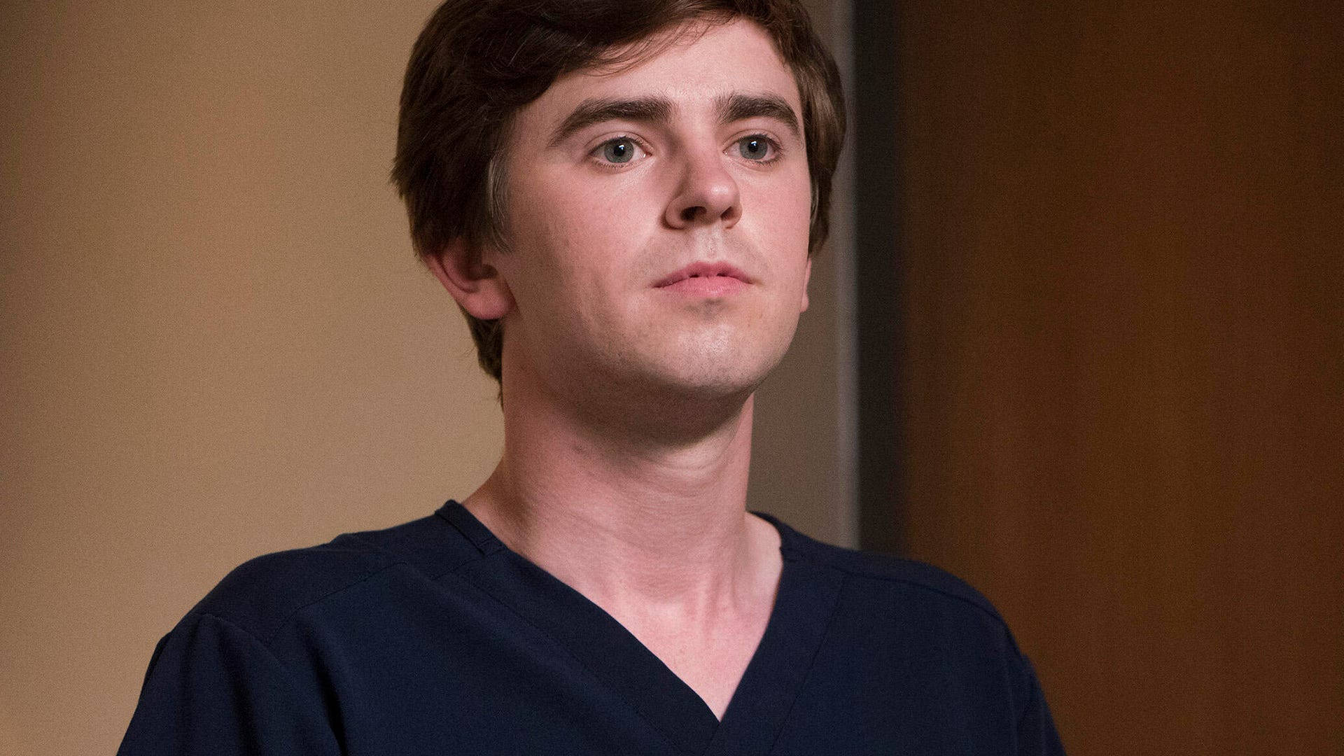 Freddie Highmore 桌布 壁紙 — 一名身穿深藍色手術服的年輕男子，在溫暖的米色和棕色背景前沉思地向上凝視。