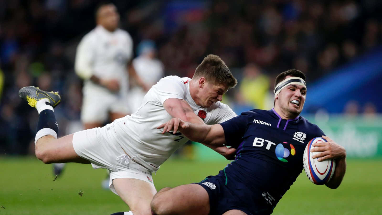Scotland Rugby 桌布 壁紙 — 一名身穿深藍色球衣的橄欖球運動員在夜間比賽中緊緊抓住球，同時被一名身穿白色球衣的對手擒抱。