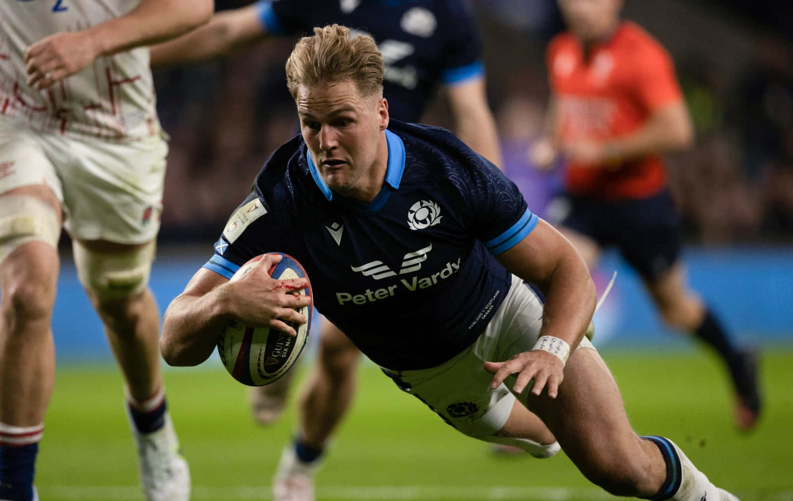 Scotland Rugby 桌布 壁紙 — 一名身穿深藍色球衣的橄欖球運動員在體育場燈光下進行激烈比賽時，抱著球向前飛撲。