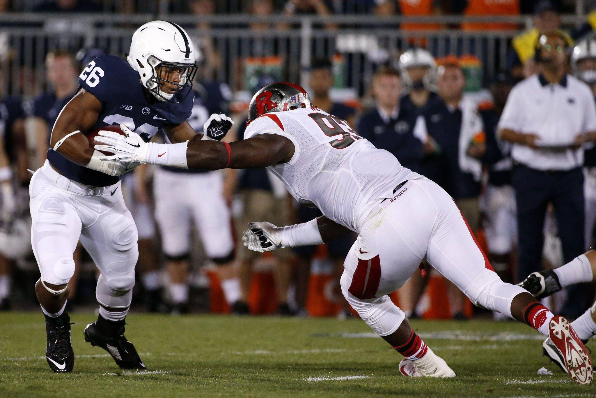 Saquon Barkley 桌布 壁紙 — 一名身穿深藍色球衣的足球員帶著球奔跑，一名身穿白色球衣的防守球員在燈光球場上攔截他。