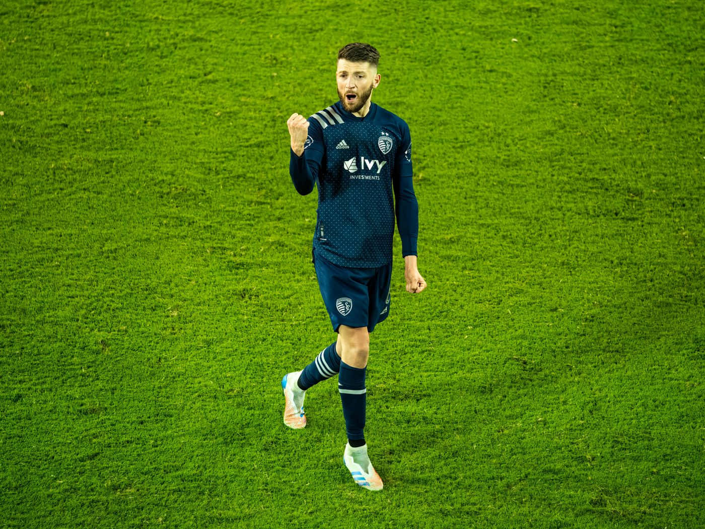 Football Players 桌布 壁紙 — 一名身穿深藍色球衣的足球運動員在體育場燈光下，於充滿活力的綠色球場上舉起拳頭慶祝。