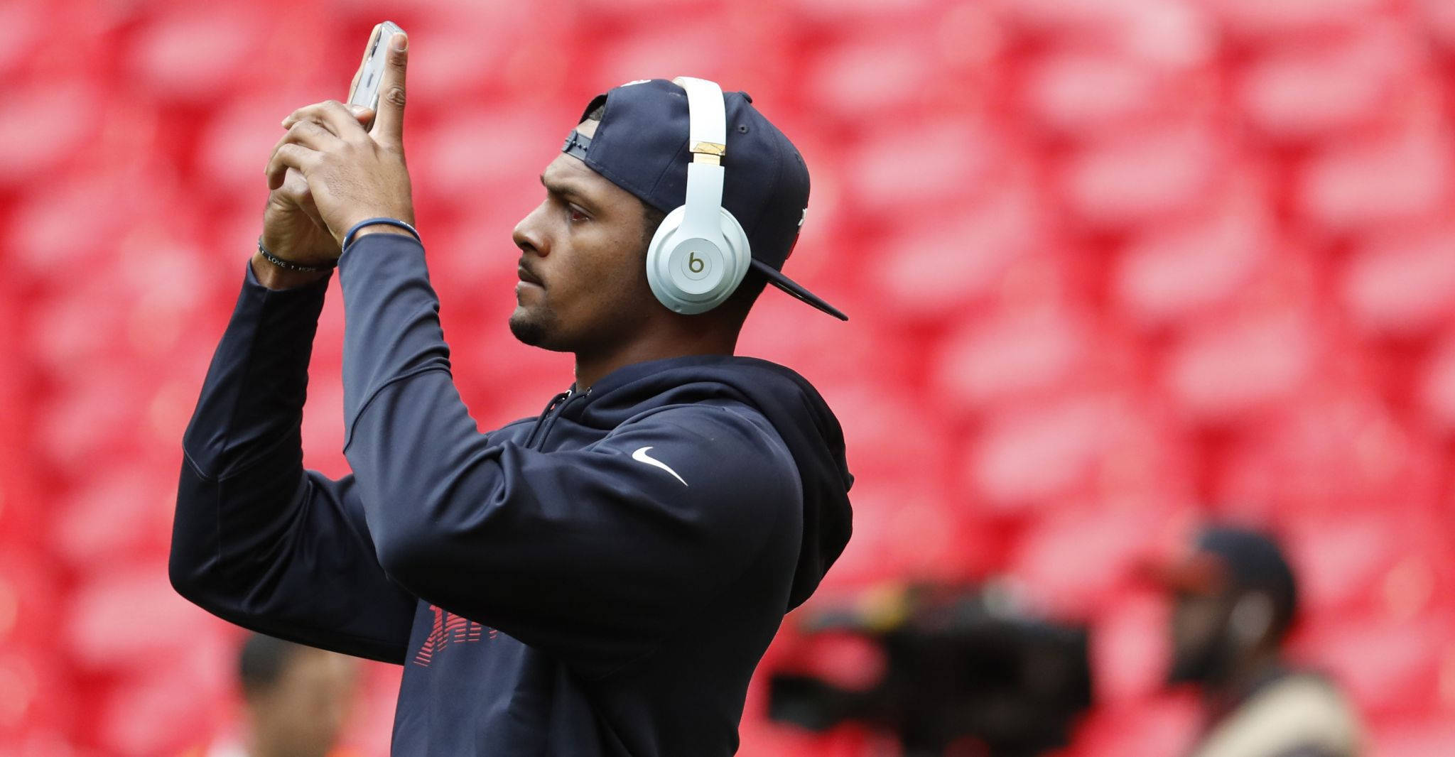 Deshaun Watson 桌布 壁紙 — 一名身穿深藍色連帽衫和白色耳機的男子，以模糊的紅色體育場座位為背景自拍。