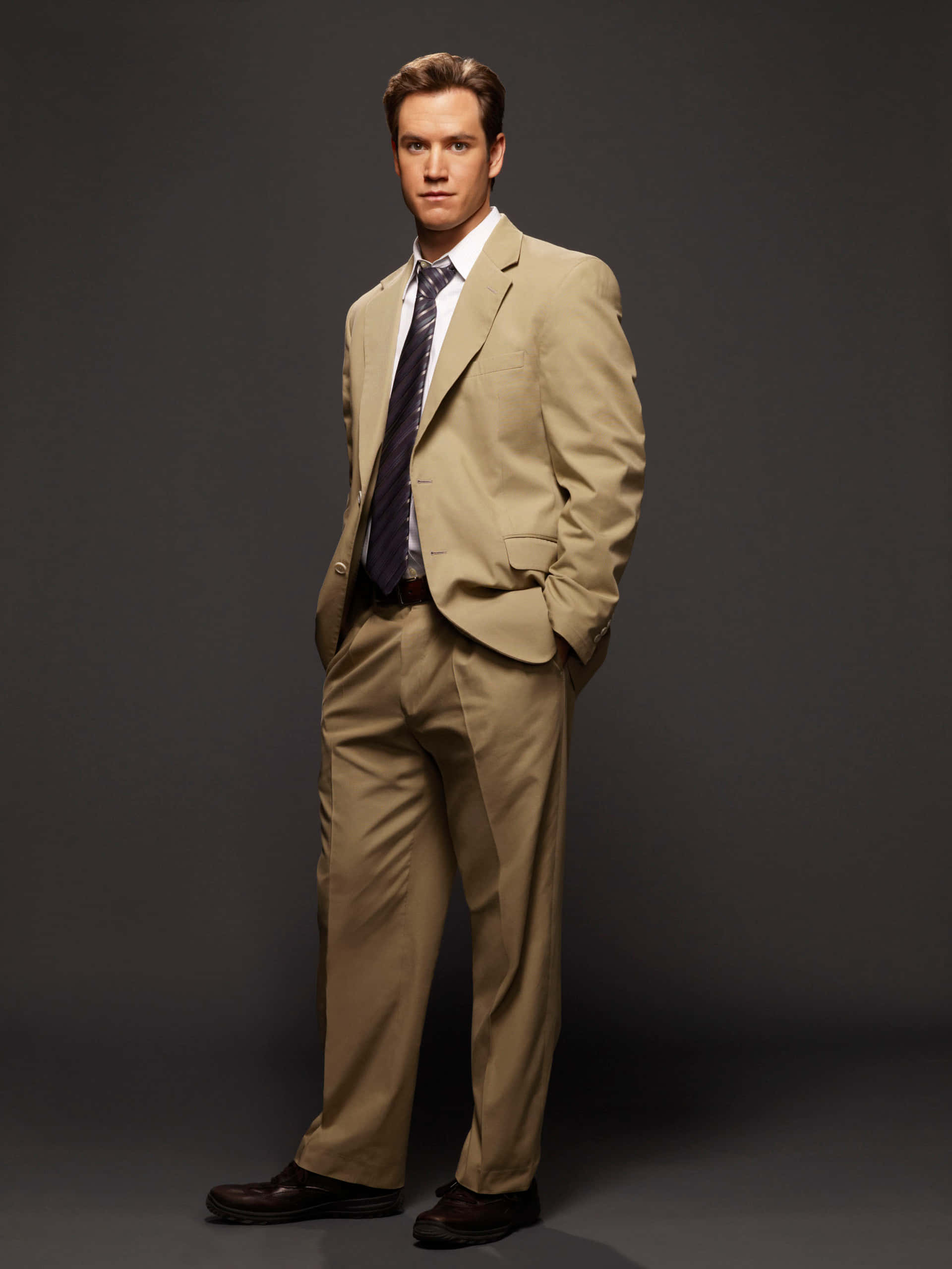 Mark-paul Gosselaar 桌布 壁紙 — 一名身穿淺棕色西裝的男子，雙手插袋，自信地站在深灰色攝影棚背景前。