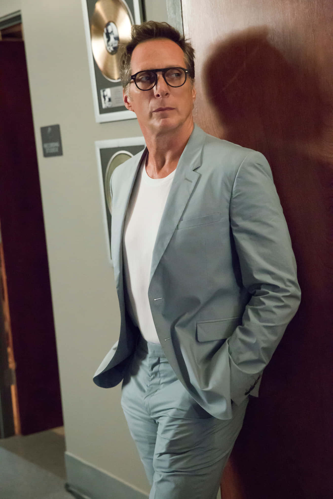 William Fichtner 桌布 壁紙 — 一名身穿淺灰色西裝、戴眼鏡的男子在走廊裡靠著牆，旁邊有鑲框的金唱片。