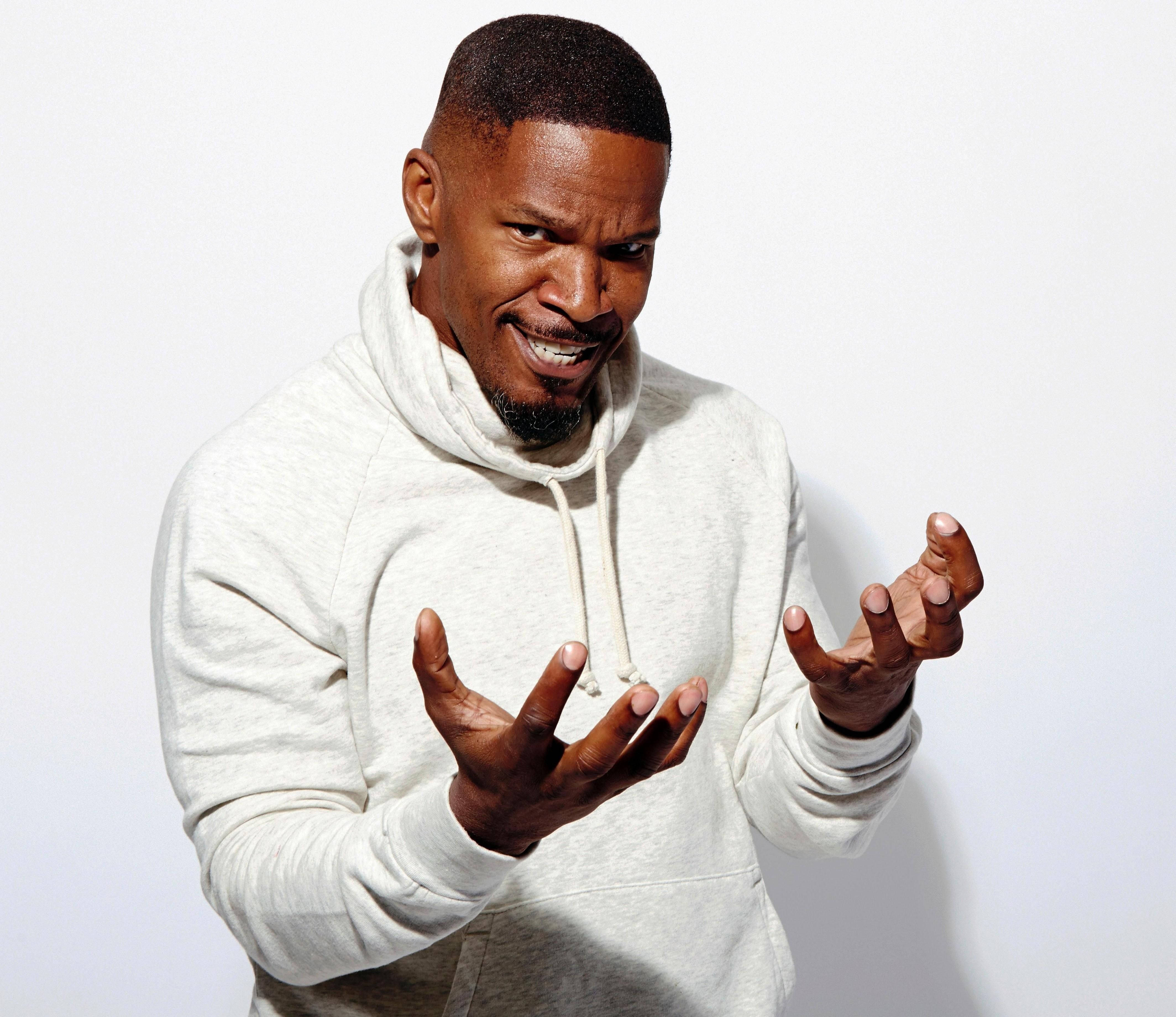 Jamie Foxx 桌布 壁紙 — 一名身穿淺灰色連帽衫的男子在純白色背景前做出一個俏皮的聳肩手勢。