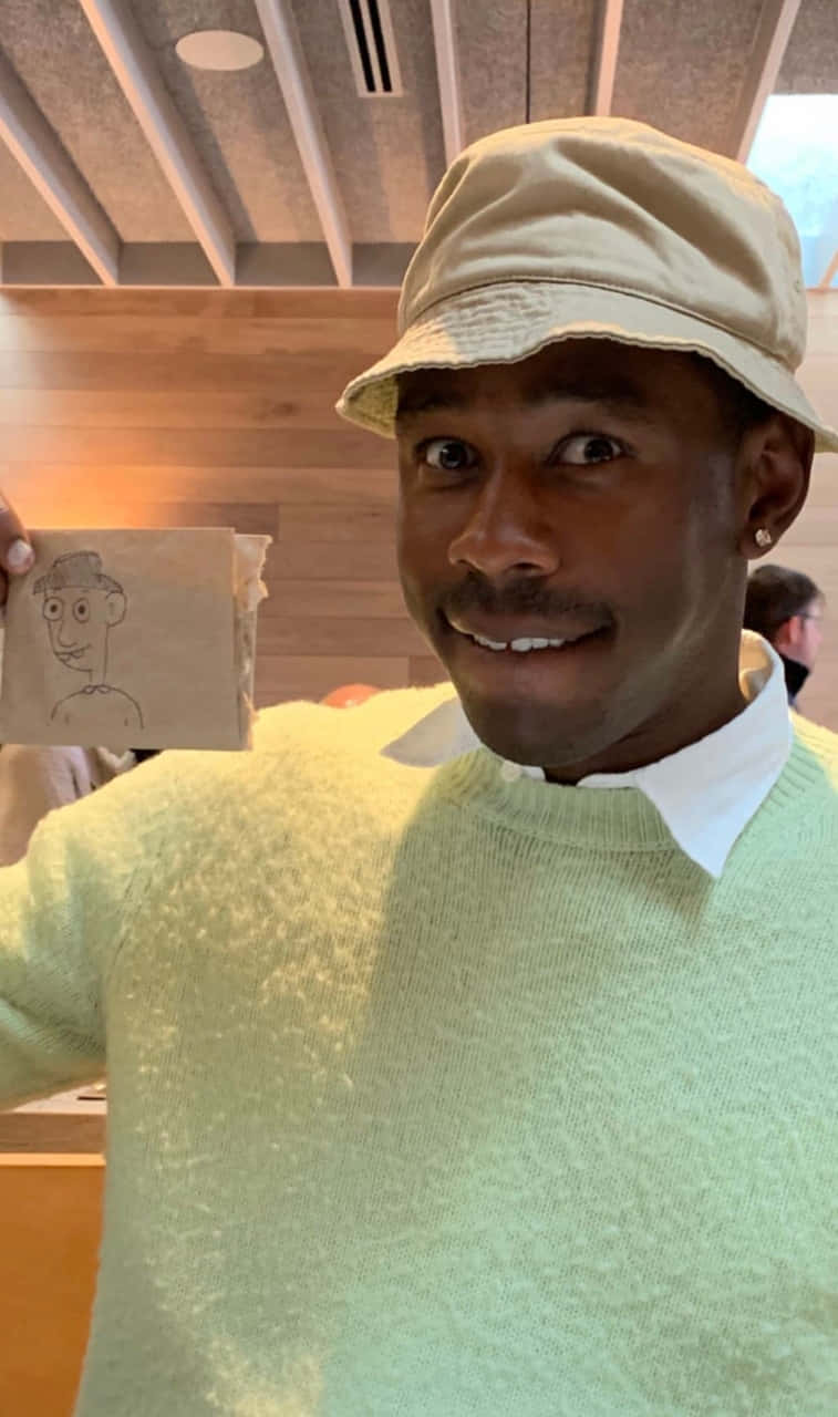 Rappers 桌布 壁紙 — 一名身穿淺綠色毛衣、戴著米色帽子的男子微笑著舉起一張簡單的鉛筆畫人臉。