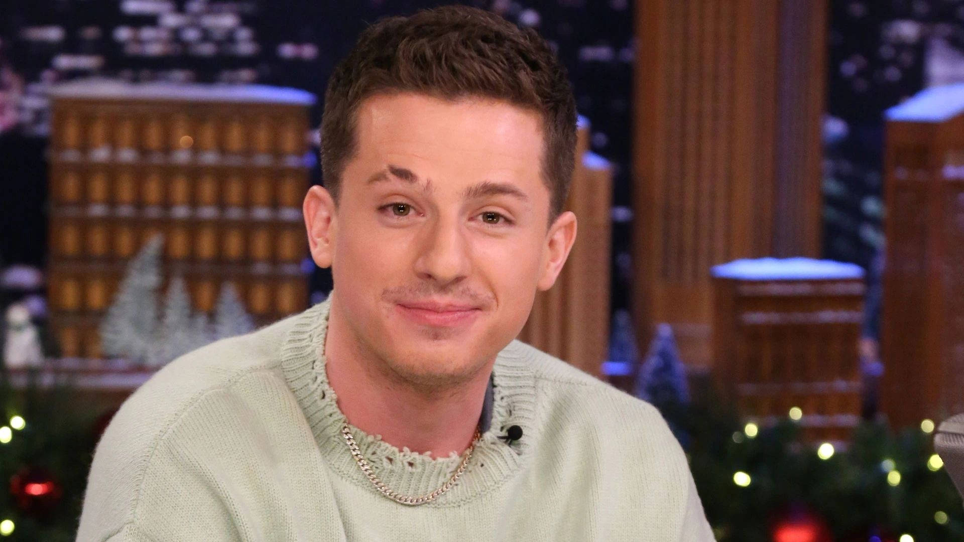 Charlie Puth 桌布 壁紙 — 一名身穿淺綠色毛衣的男子在節日氣氛濃厚的脫口秀舞台上微笑，背景是雪景城市天際線和閃爍的燈光。