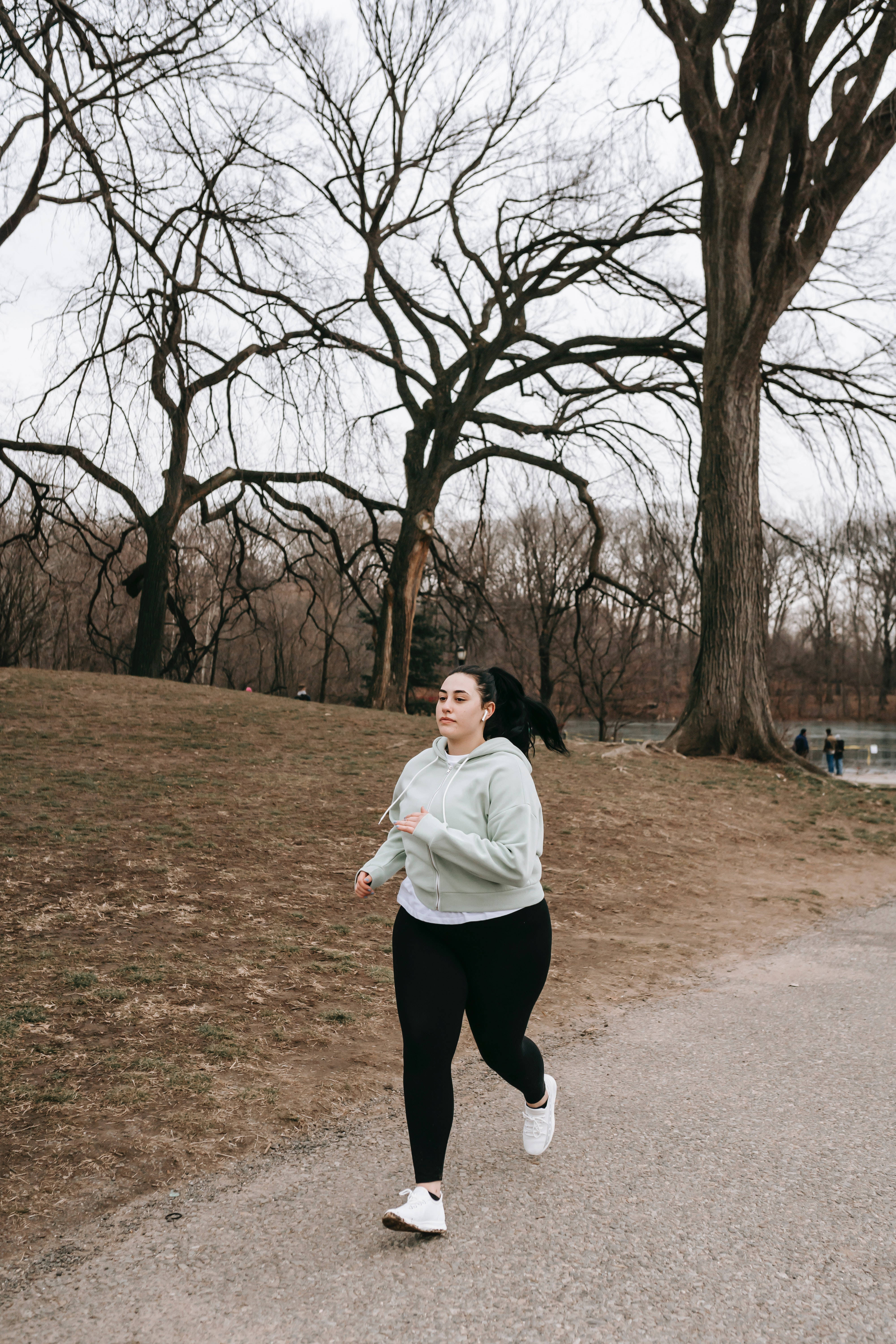 Fitness 桌布 壁紙 — 一名身穿淺綠色連帽衫的女子在陰天之下，沿著公園小徑奔跑，兩旁是光禿禿的冬季樹木。