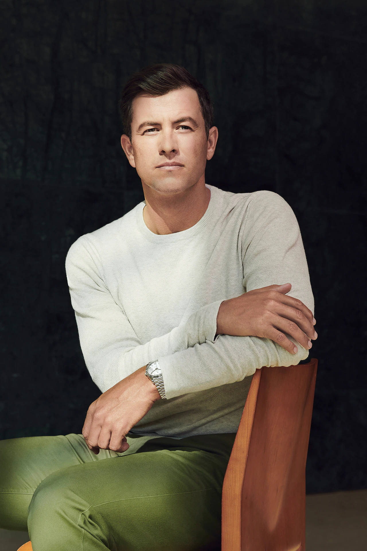 adam scott 桌布 壁紙 — 一名身穿淺色毛衣和綠色褲子的男子自信地坐在木椅上，背景是深色紋理。