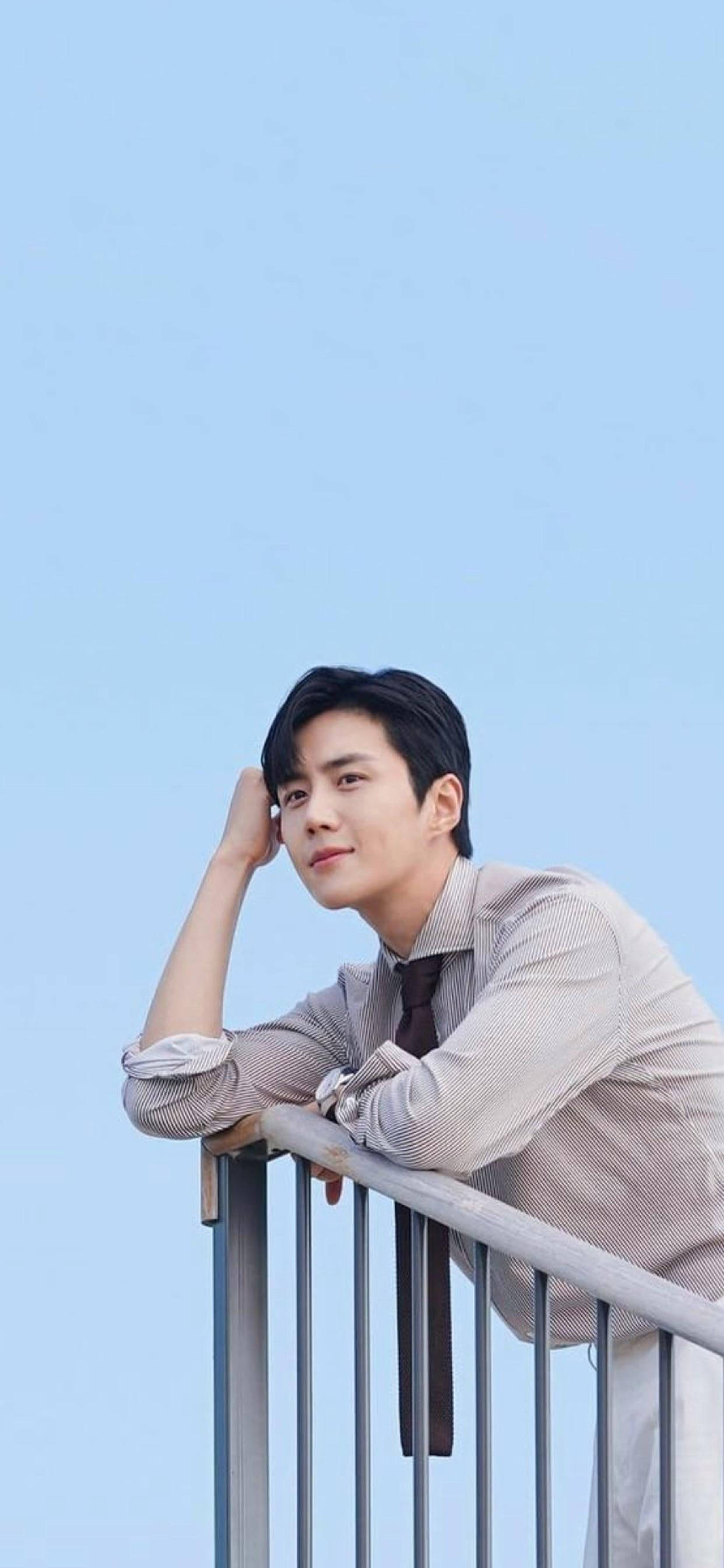 Kim Seon Ho 桌布 壁紙 — 一名身穿淺色襯衫的年輕男子，在清澈的藍天下若有所思地靠在欄杆上。