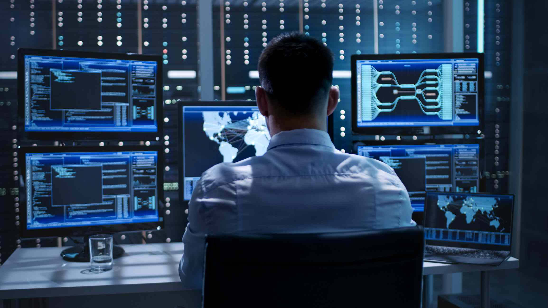 Working 桌布 壁紙 — 一名身穿淺色襯衫的技術員坐在辦公桌前，被顯示程式碼和世界地圖的藍色發光顯示器包圍，身處昏暗的伺服器機房。
