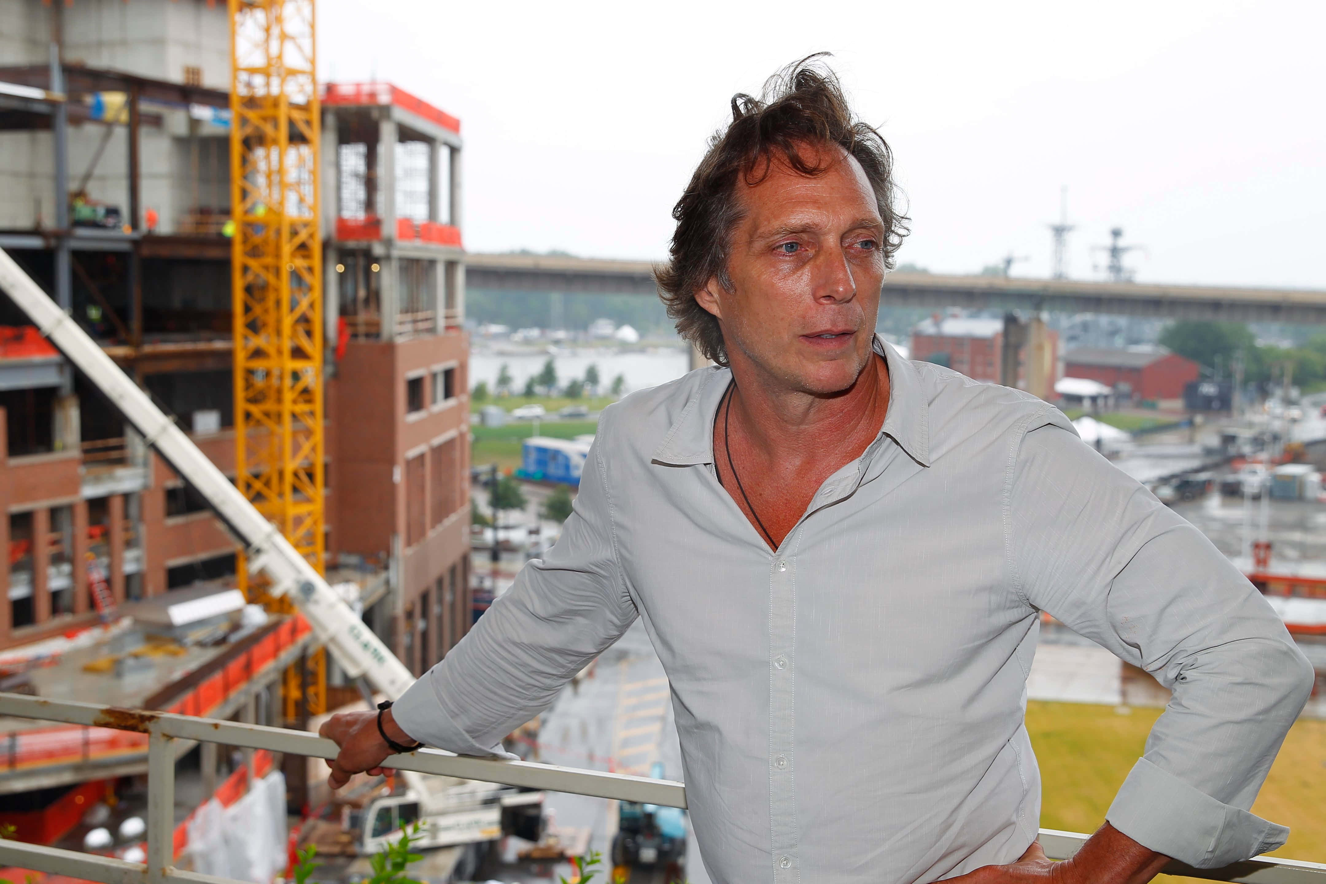 William Fichtner 桌布 壁紙 — 一名身穿淺色襯衫的男子靠在欄杆上，俯瞰著一個繁忙的建築工地，背景有起重機和港口。