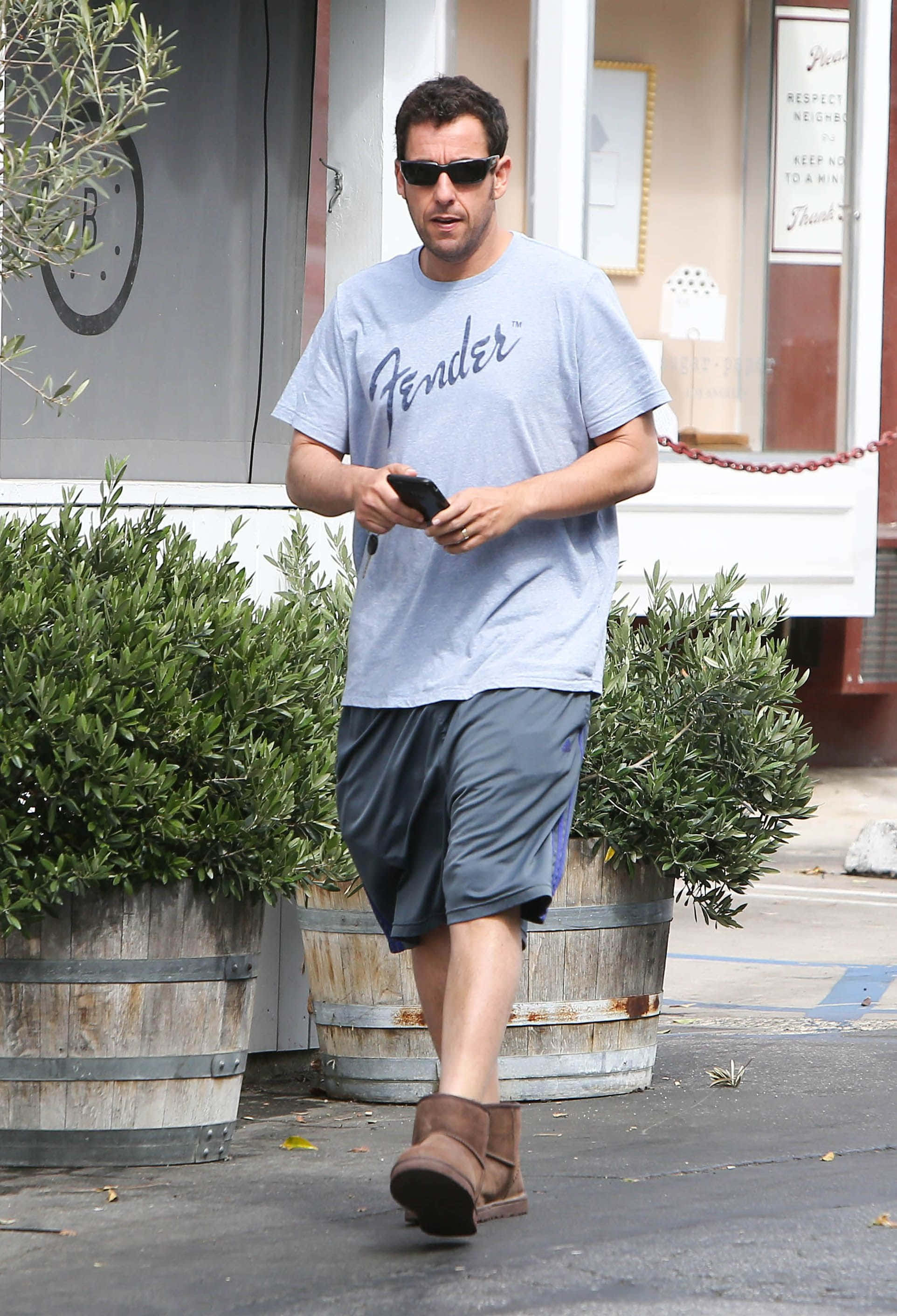 Adam Sandler 桌布 壁紙 — 一名身穿淺藍色Fender T恤和灰色短褲的男子，一邊看著手機一邊在戶外行走。