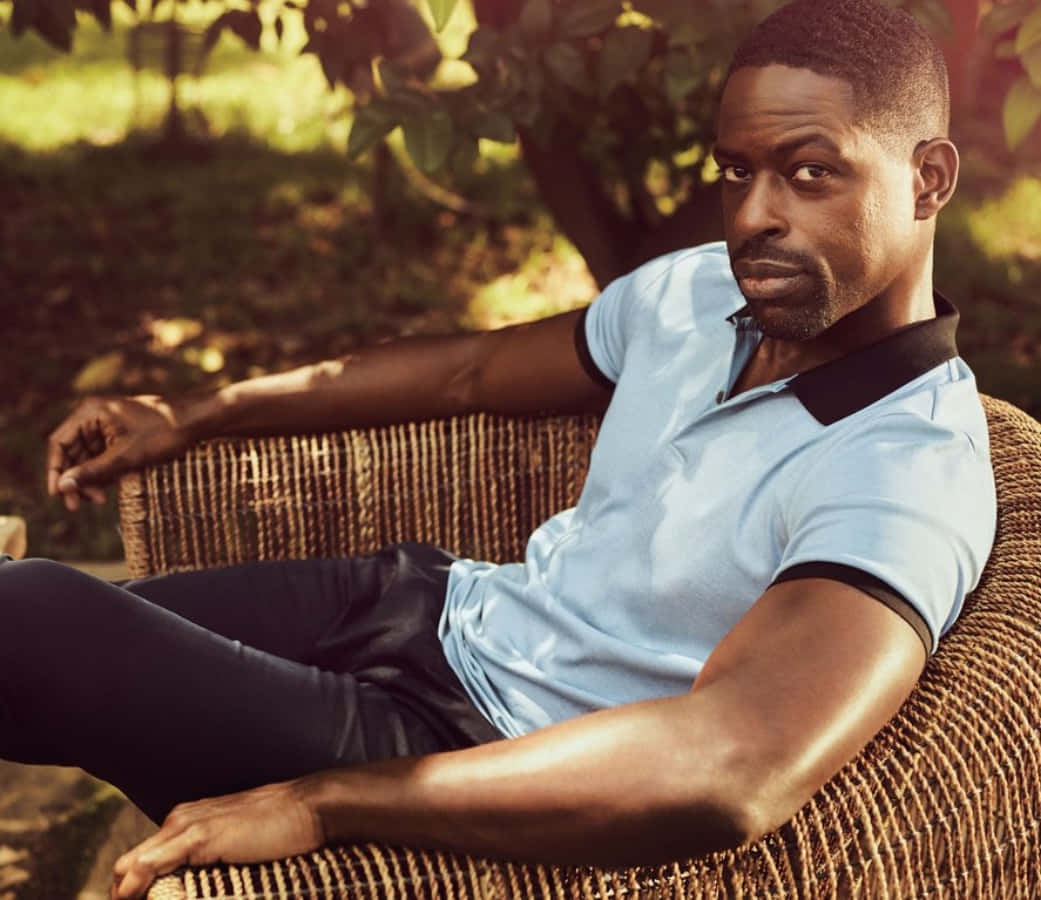 Sterling K Brown 桌布 壁紙 — 一名身穿淺藍色Polo衫的男子，在茂密花園的斑駁陽光下，自信地靠在藤椅上。
