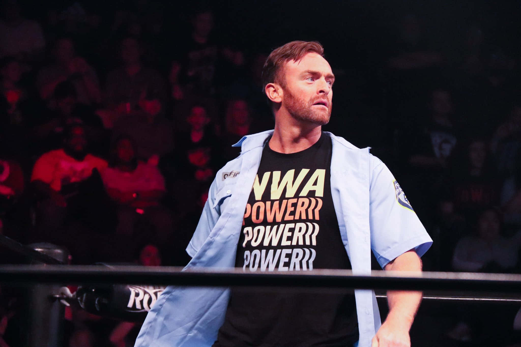 Nwa 桌布 壁紙 — 一名身穿淺藍色外套和黑色「NWA POWER!!」T恤的摔角手站在擂台上，身後有群眾。