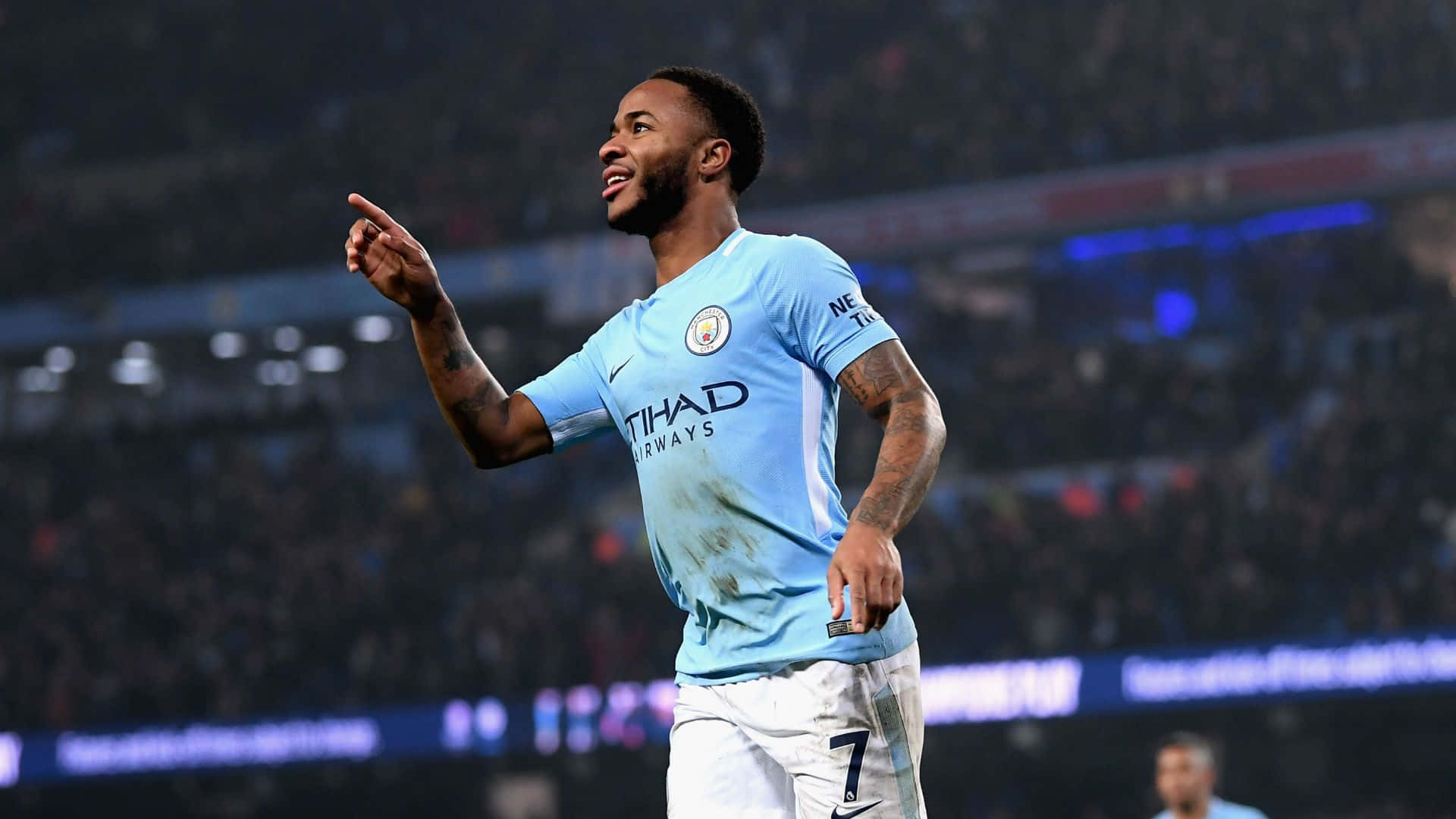 Sterling 桌布 壁紙 — 一名身穿淺藍色曼城球衣的足球員在夜晚模糊的球場人群中舉起手指慶祝。