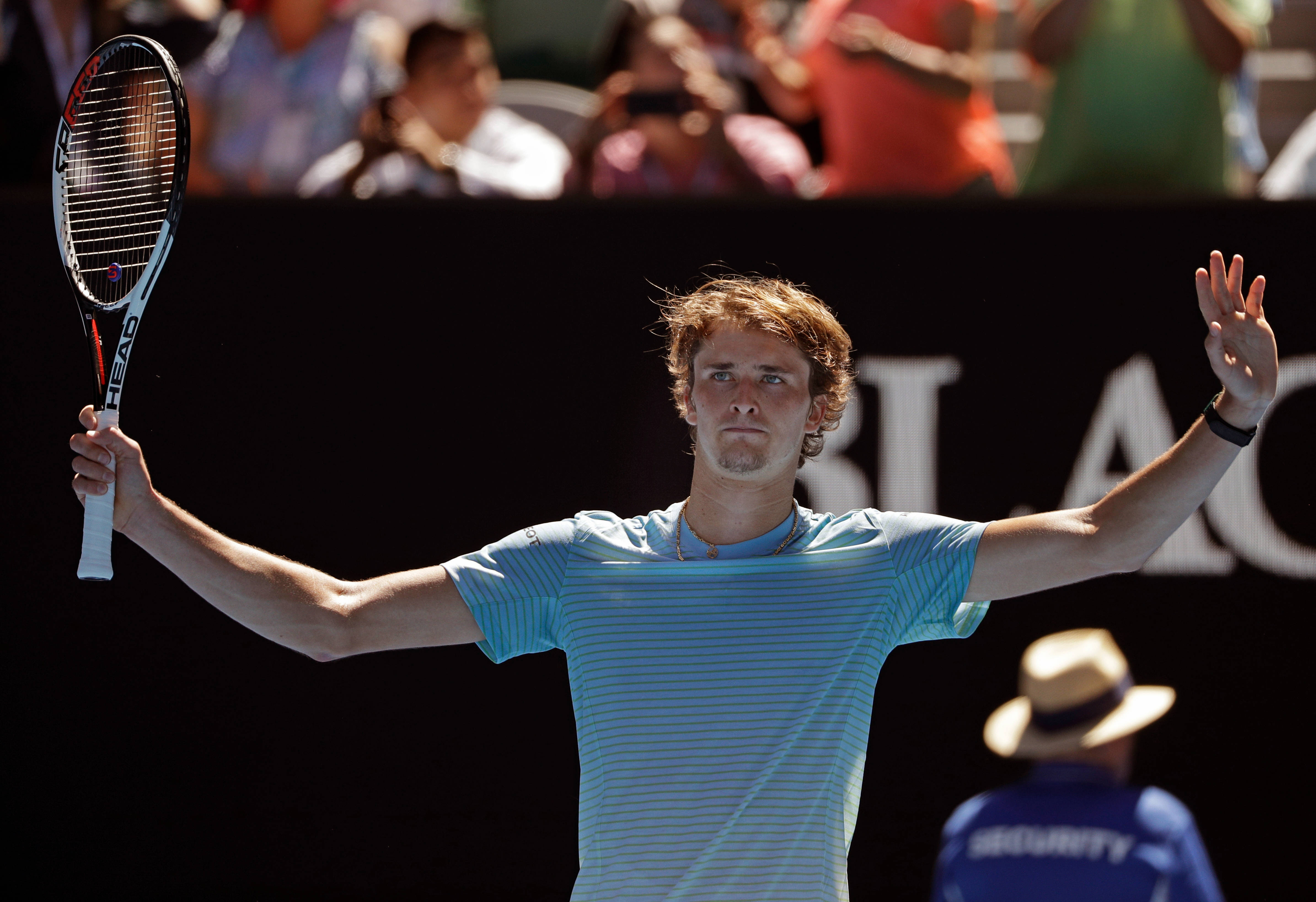 alexander zverev 桌布 壁紙 — 一名身穿淺藍色條紋襯衫的男網球運動員在明亮的日光下拿著球拍向人群揮手。