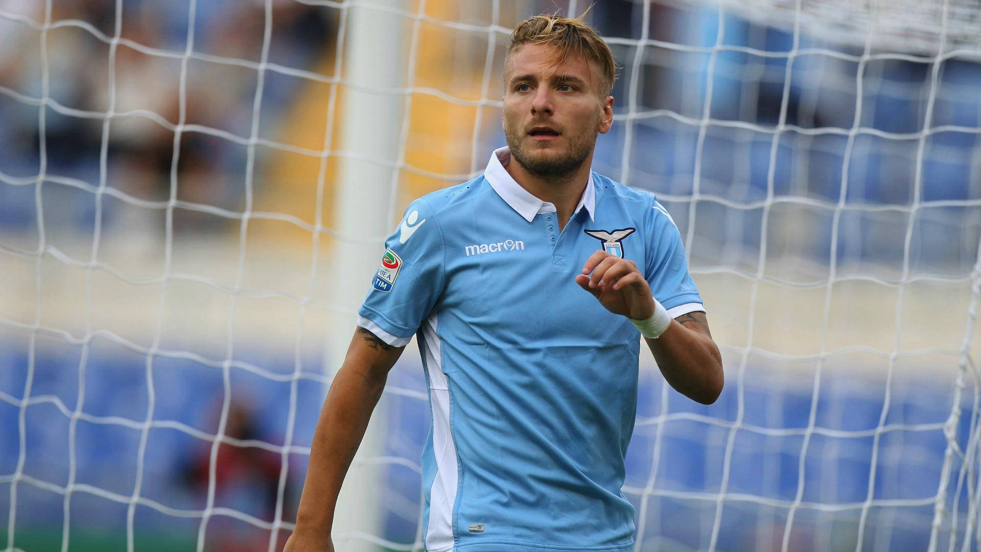 ciro immobile 桌布 壁紙 — 一名身穿淺藍色球衣的足球員站在球門網附近，背景是模糊的體育場座位。