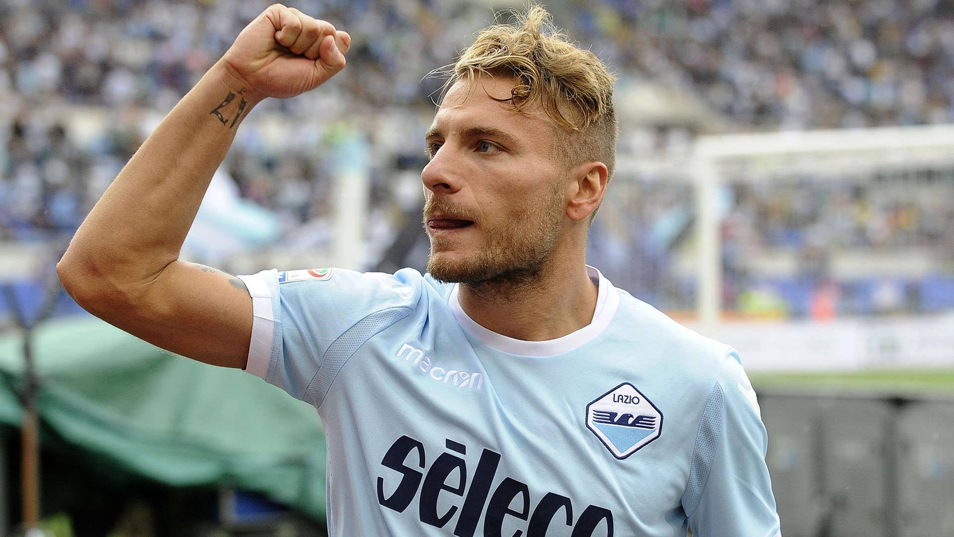 ciro immobile 桌布 壁紙 — 一名身穿淺藍色球衣的足球運動員在陽光明媚的球場上，身後有模糊的觀眾，勝利地舉起拳頭。