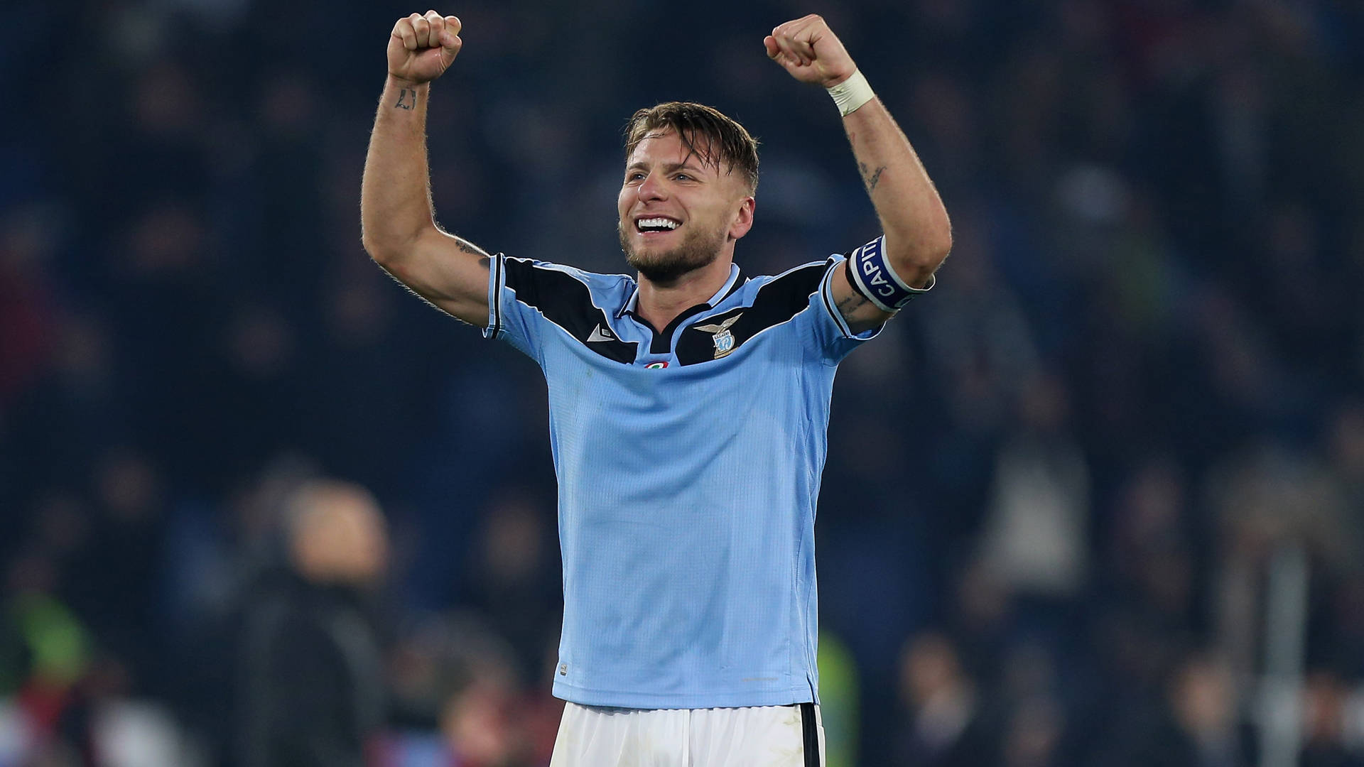 ciro immobile 桌布 壁紙 — 一名身穿淺藍色球衣的足球運動員在體育場燈光下高舉雙拳，露出勝利的燦爛笑容。