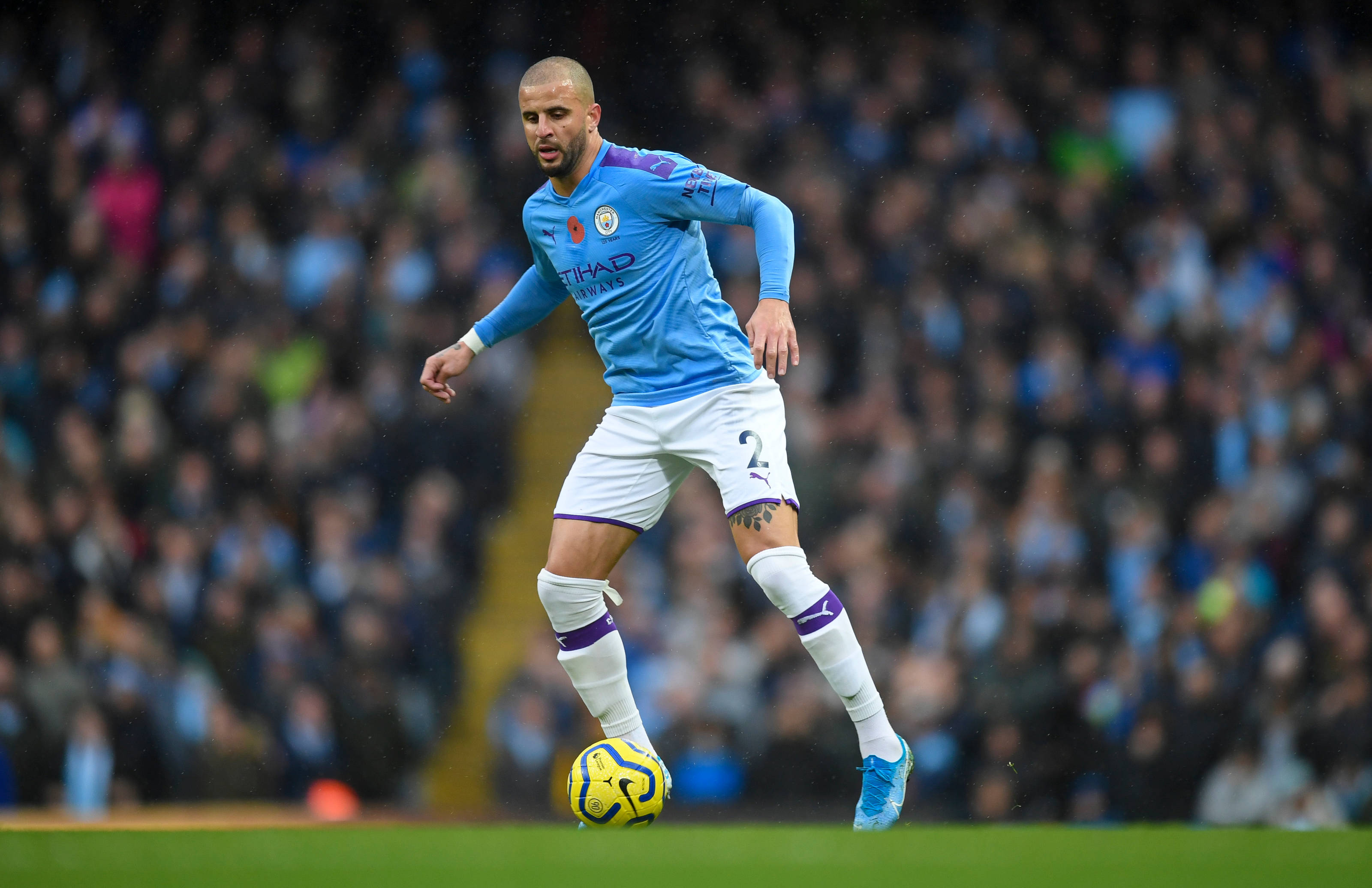 Kyle Walker 桌布 壁紙 — 一名身穿淺藍色球衣的足球運動員在綠色球場上控制著一個黃色足球，背景是模糊的人群。