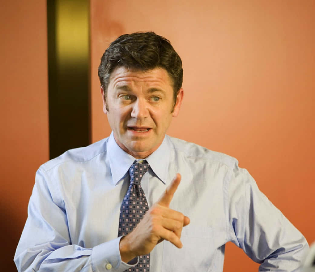 John Michael Higgins 桌布 壁紙 — 一名身穿淺藍色襯衫和圖案領帶的男子，在溫暖的橙色牆壁前，表情豐富地向前指。