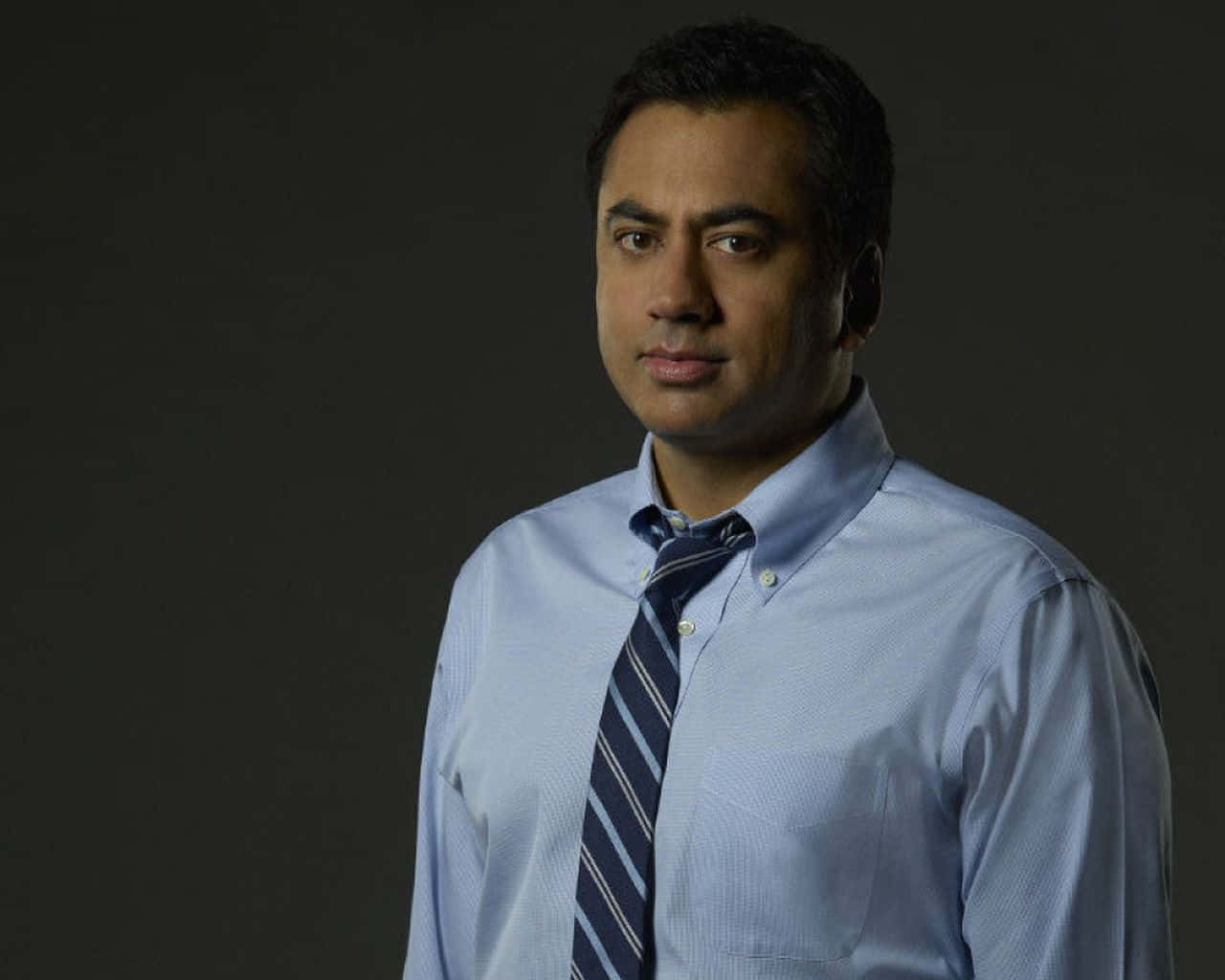Kal Penn 桌布 壁紙 — 一名身穿淺藍色襯衫和條紋領帶的男子站在深灰色攝影棚背景前，光線柔和。