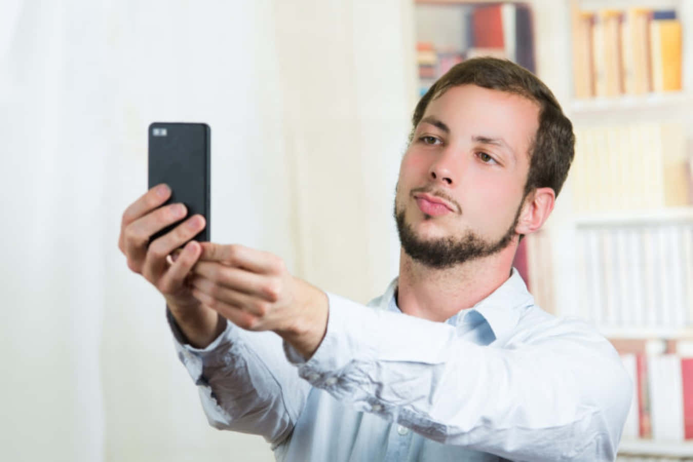 selfie 桌布 壁紙 — 一名身穿淺藍色襯衫的男子在身後有書架的房間裡用黑色手機自拍。