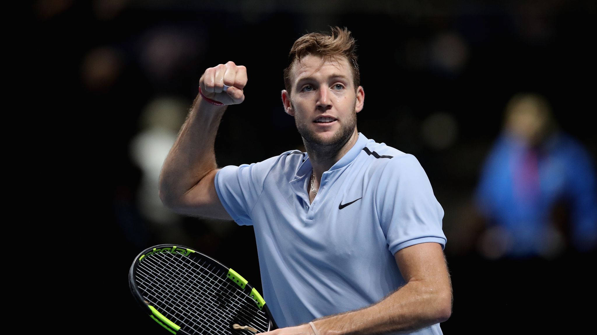 Tennis 桌布 壁紙 — 一名身穿淺藍色襯衫的男性網球運動員，在黑暗模糊的背景前，握著球拍，勝利地揮舞拳頭。