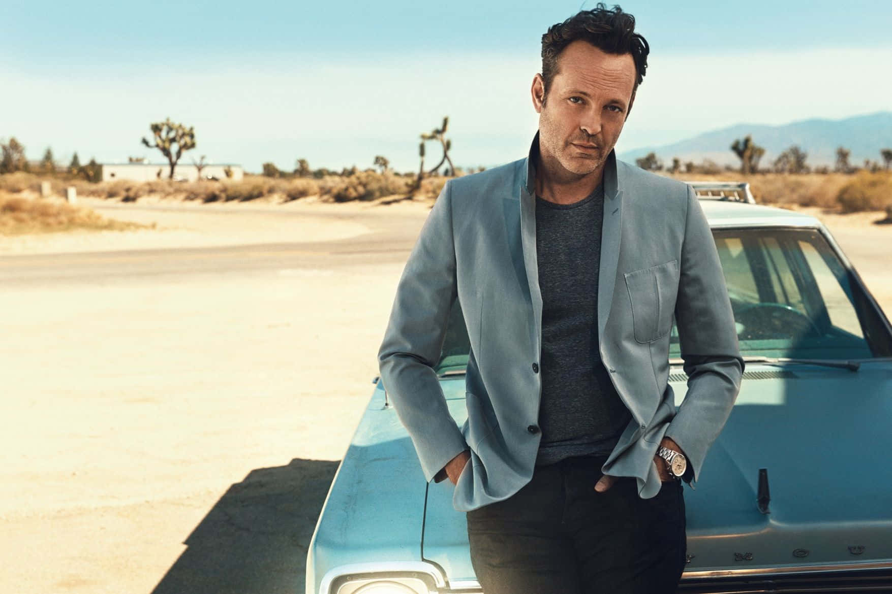 Vince Vaughn 桌布 壁紙 — 一名身穿淺藍色西裝外套的男子在陽光明媚的沙漠中，倚靠著一輛復古青綠色汽車，背景有約書亞樹和遠山。
