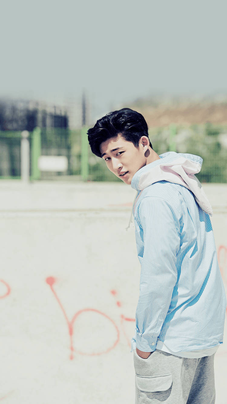 kim hanbin 桌布 壁紙 — 一名身穿淺藍色連帽衫和灰色運動褲的年輕男子站在陽光明媚的城市滑板公園中，混凝土上有塗鴉。