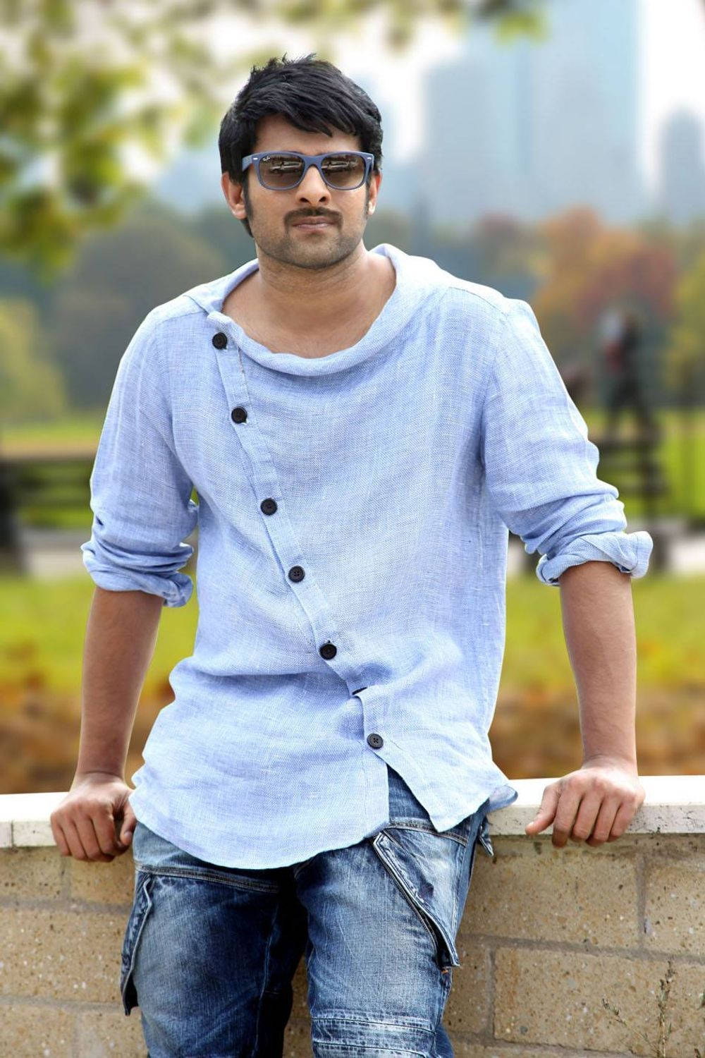 Prabhas Mirchi 桌布 壁紙 — 一名身穿淺藍色鈕扣襯衫和牛仔褲的男子，在公園裡隨意地靠在一道石牆上，背景是模糊的綠化和城市天際線。