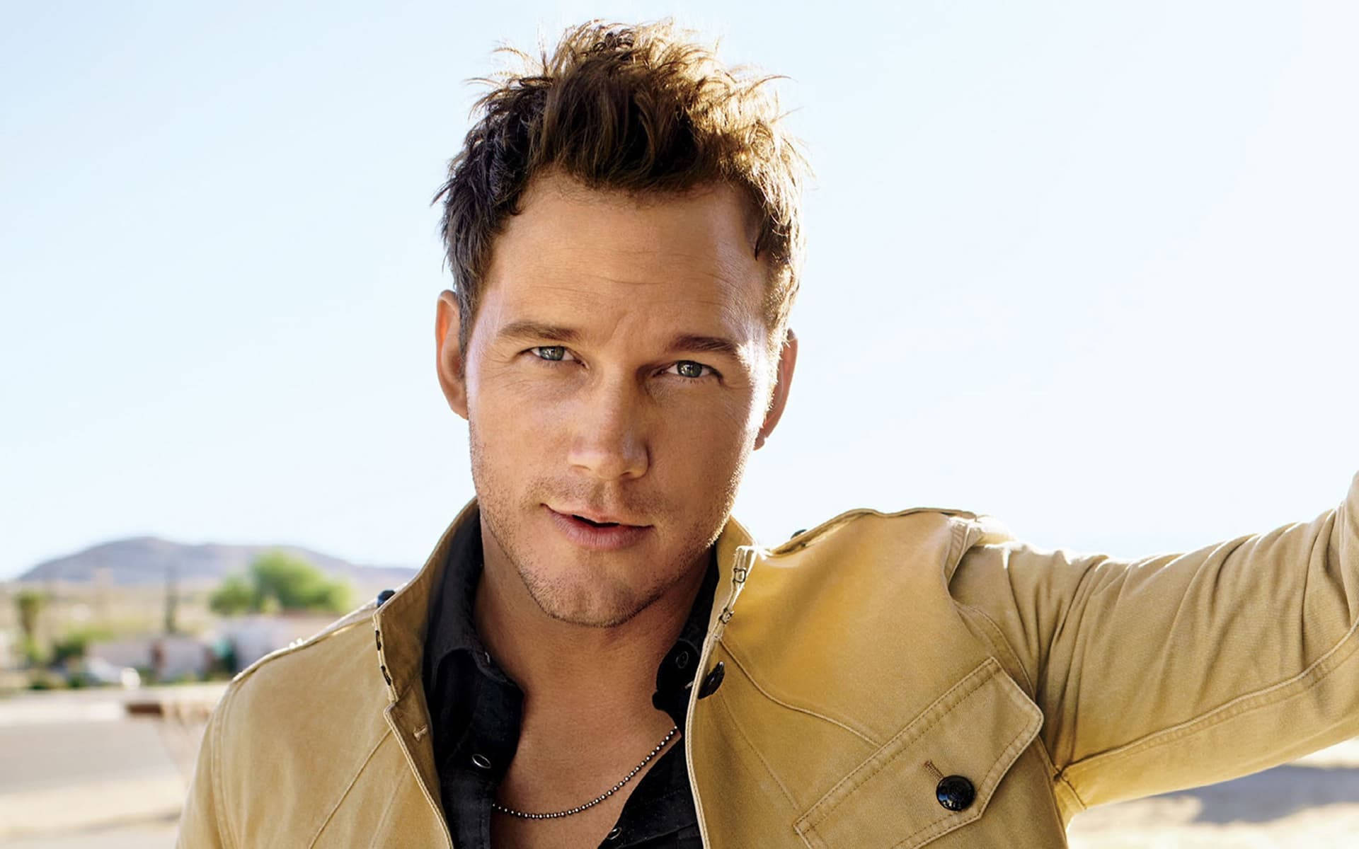 Chris Pratt 桌布 壁紙 — 一名身穿淺褐色外套的男子在晴朗天空下，以明亮的沙漠背景和遠山為襯托，自信地擺出姿勢。