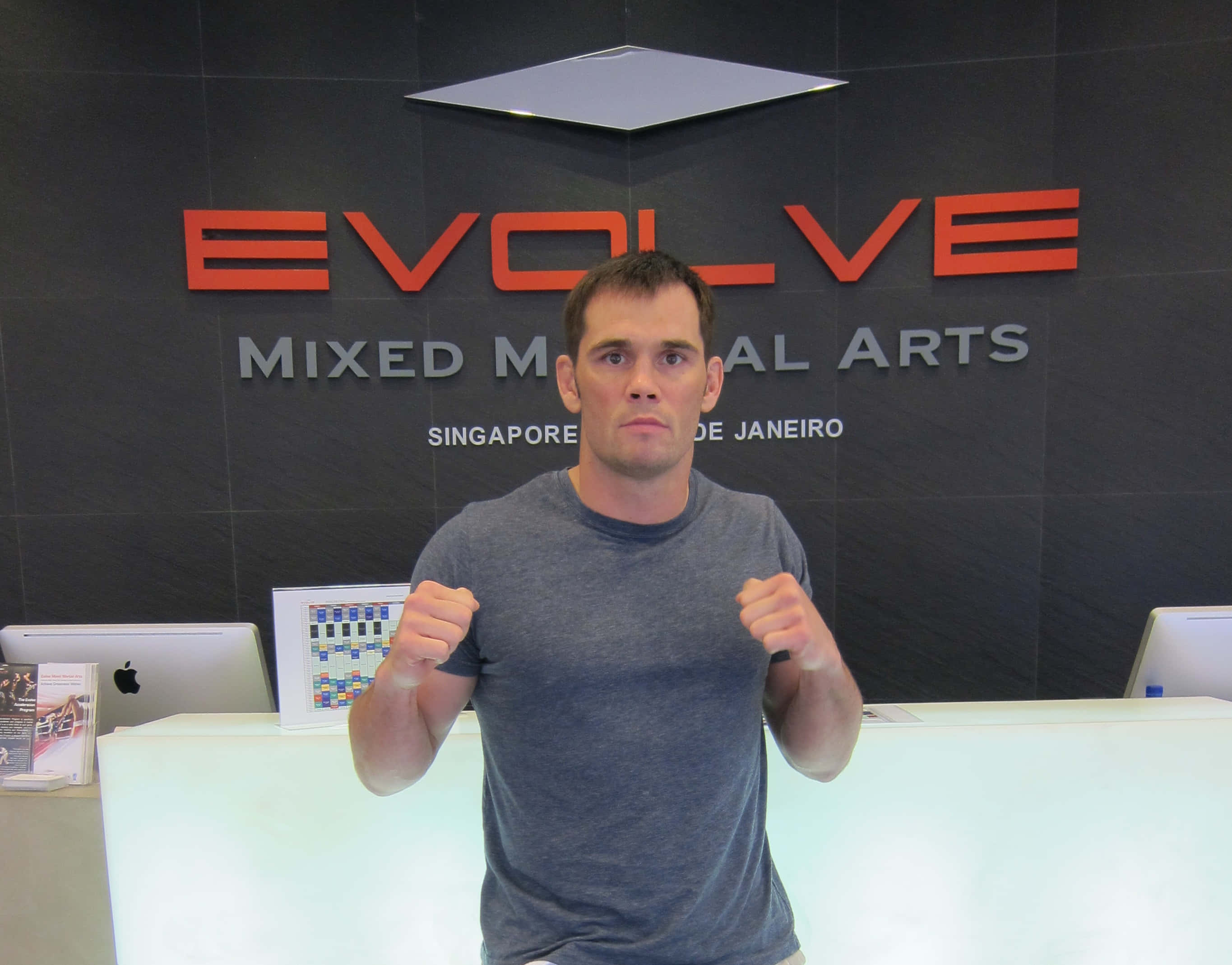 UFC 桌布 壁紙 — 一名身穿灰色T恤的男子在武術館內舉起拳頭，面向鏡頭，站在'EVOLVE'標誌下。